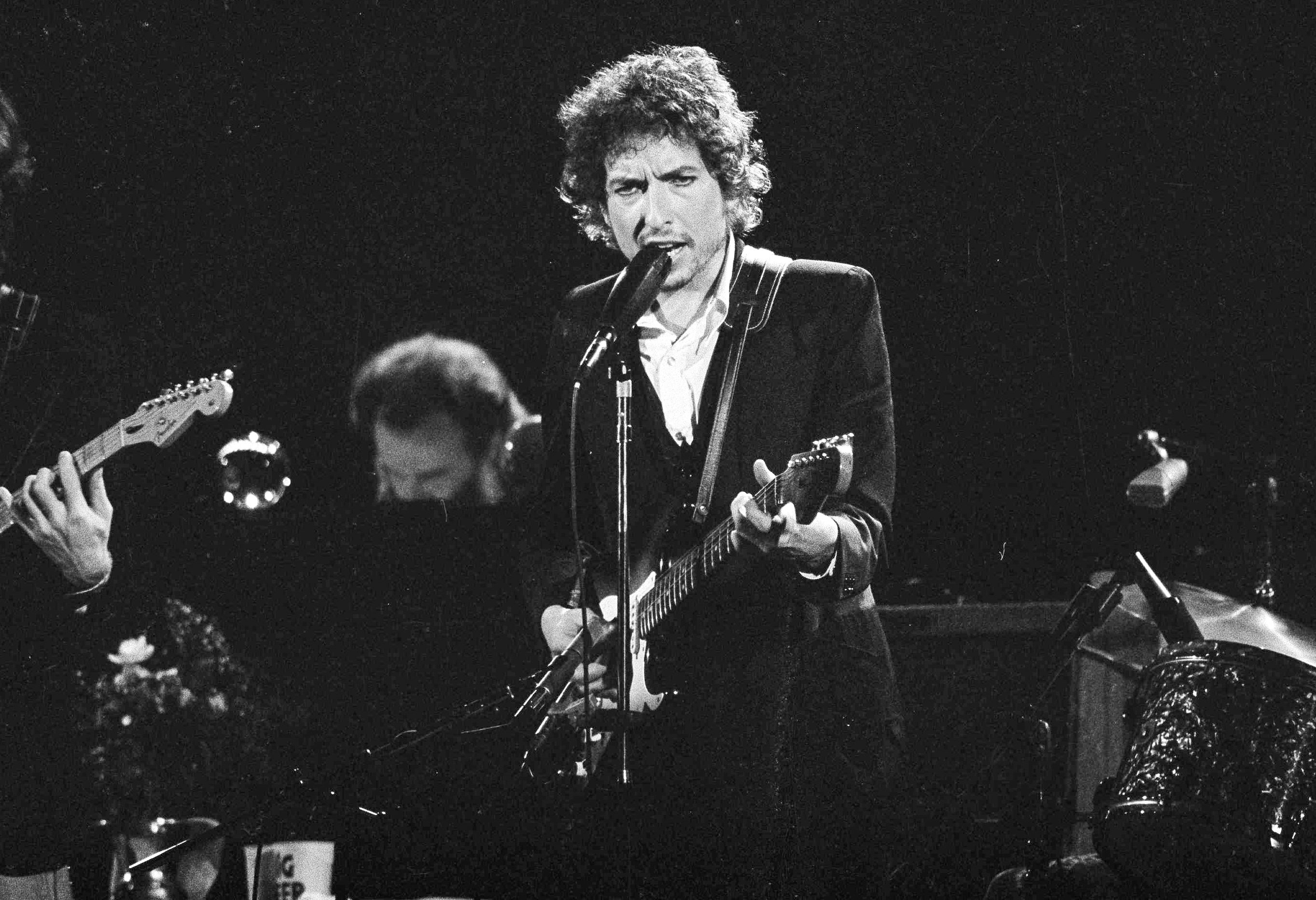 Musician Bob Dylan belts out a tune as members of The Band (Robbie Robertson, partially hidden left, and Garth Hudson, background center) accompany him, during a concert in Los Angeles on 15 February 1974