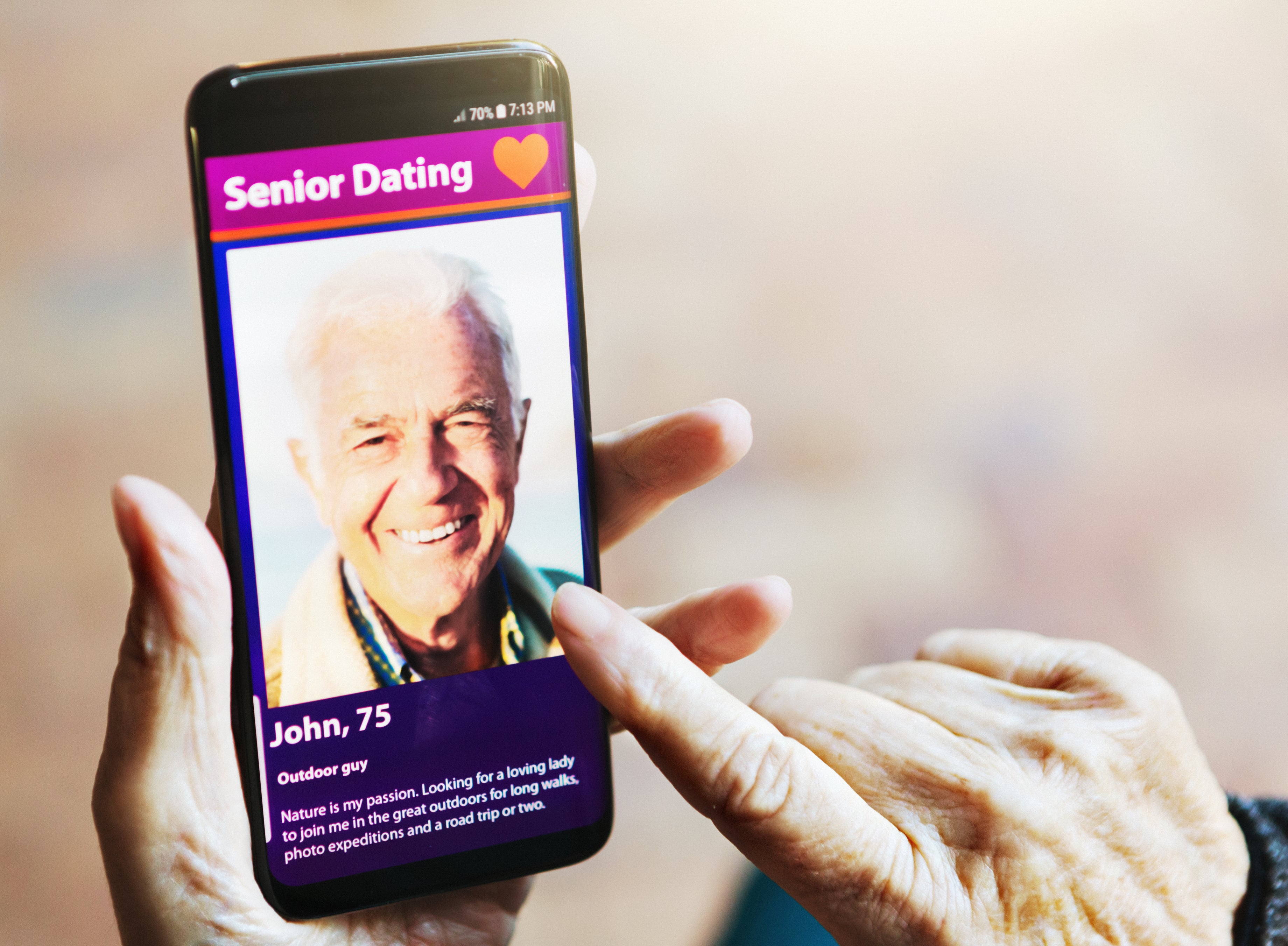 A senior woman's hand taps the screen of her phone, which shows a handsome senior man displayed on a dating app