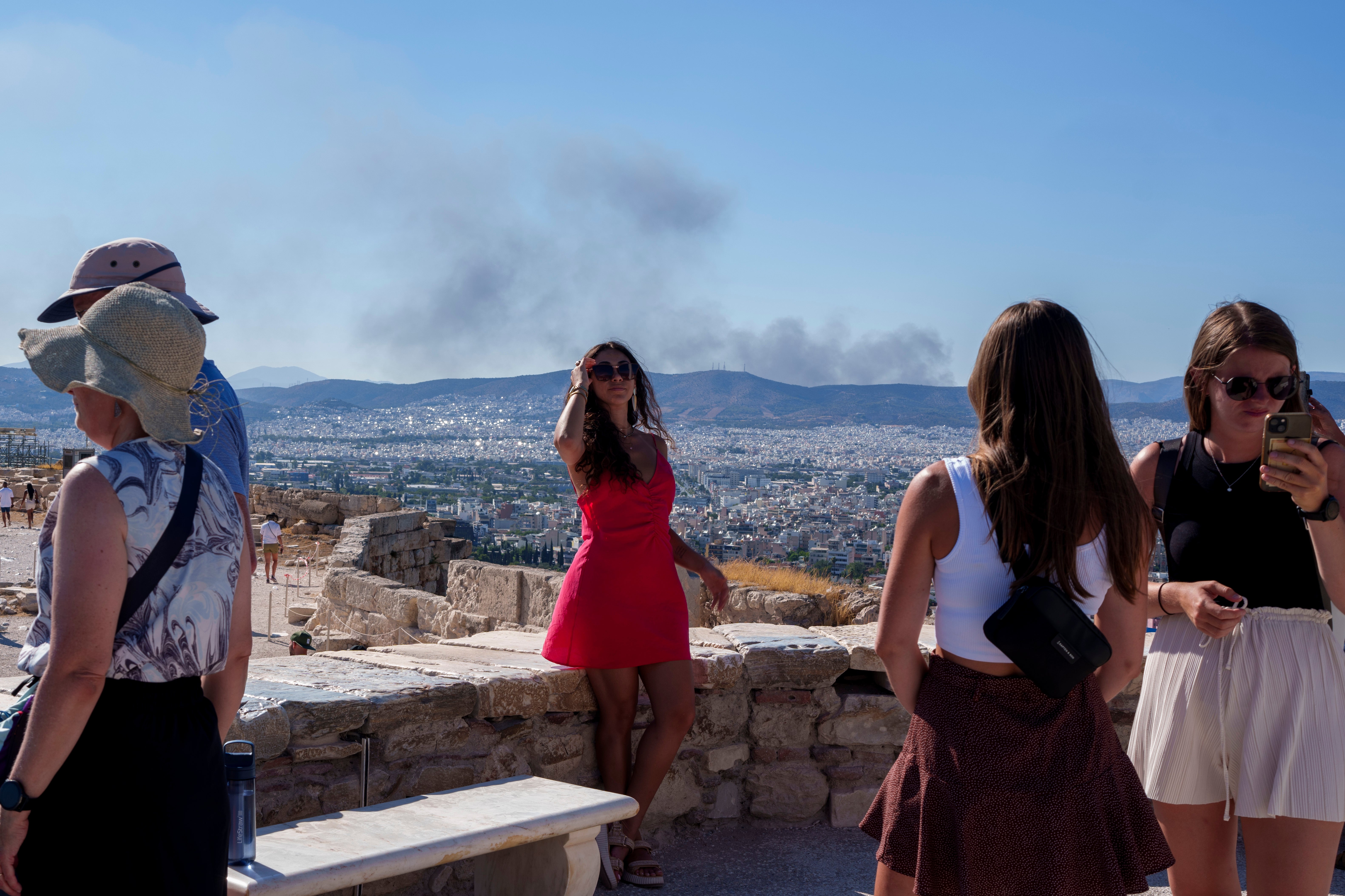 A woman poses for a photo with smoke from a fire at the background, during a hot, windy day at Acropolis hill, in Athens