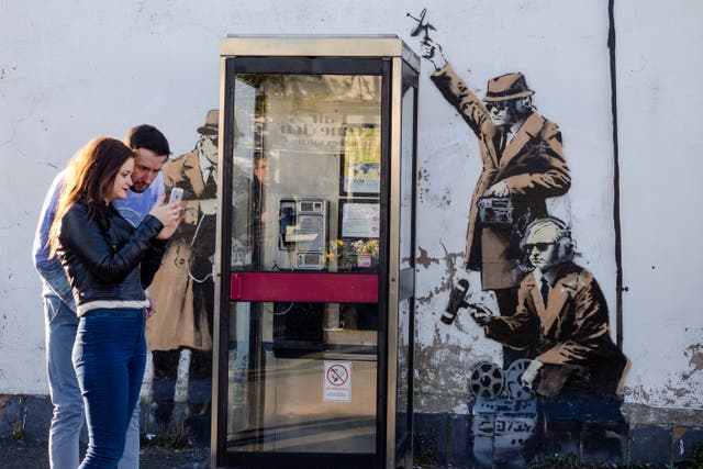 <p>Tourists in Cheltenham pose by a ‘dated’ BT phone box – complete with Banksy mural </p>
