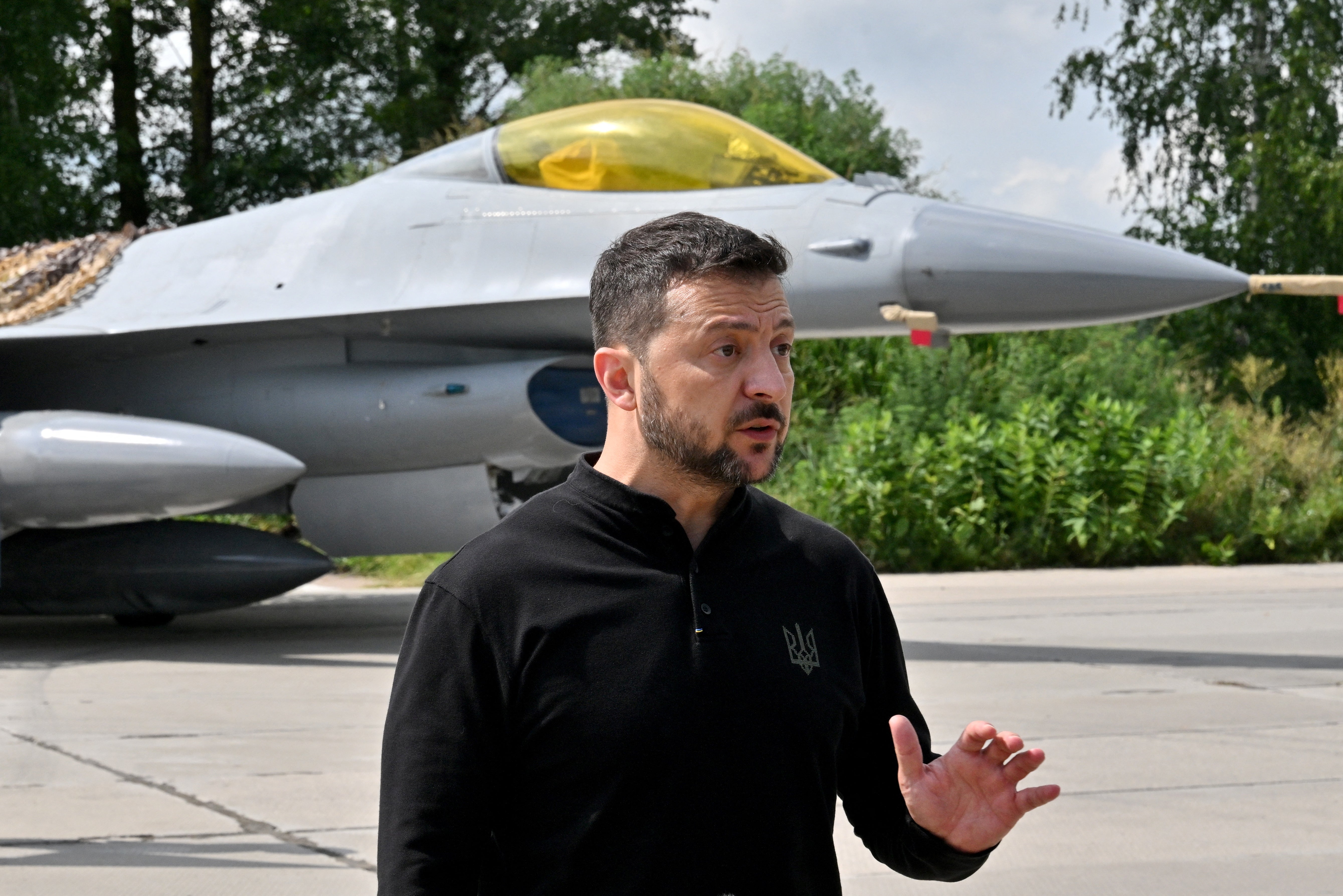 Volodymyr Zelensky gestures as he speaks to media representatives while standing in front of an F-16 fighter jet