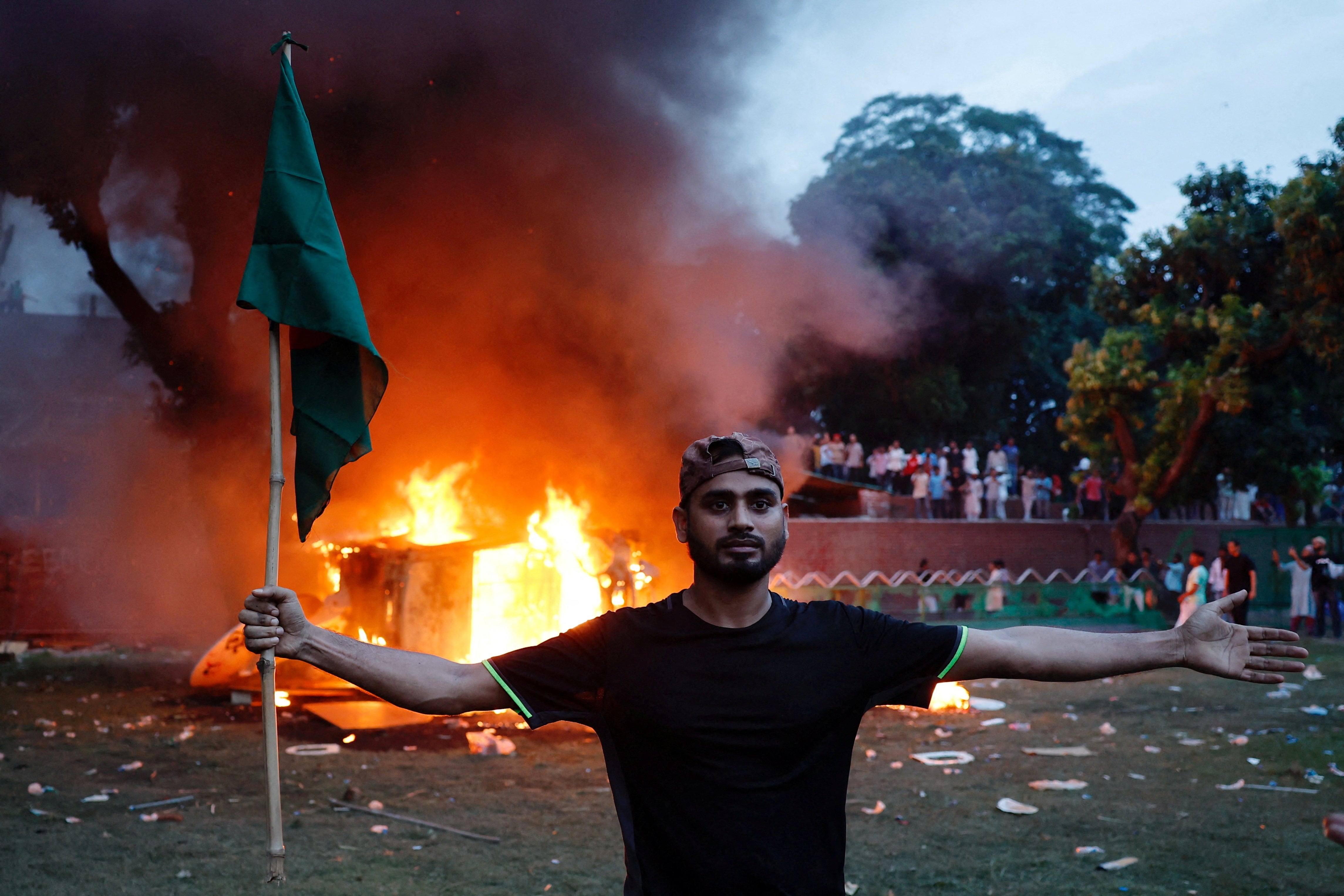 A man holding a Bangladesh flag stands in front of a vehicle that was set on fire at the Ganabhaban, the Prime Minister's residence, after the resignation of PM Sheikh Hasina in Dhaka