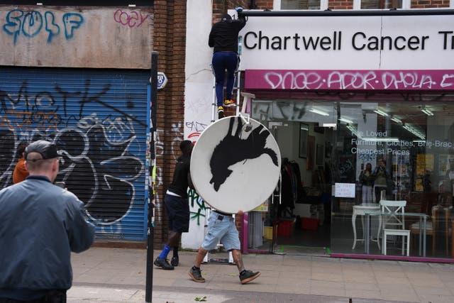 <p>People remove a new artwork by Banksy, depicting a howling wolf painted on a satellite dish that was placed on a shop roof in Peckham, south London</p>