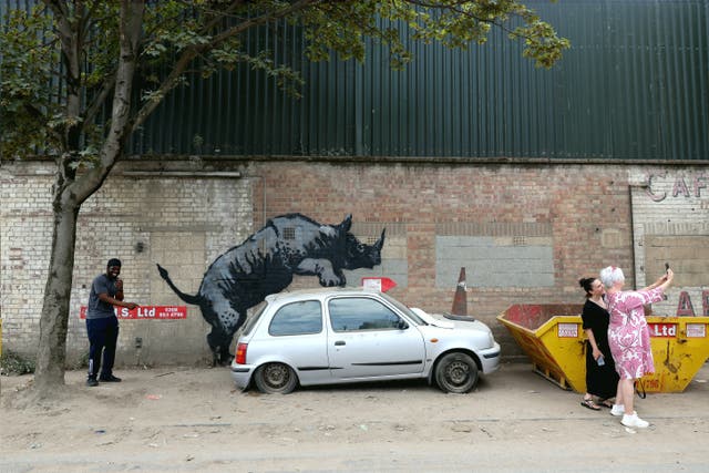 <p>People gather around an artwork by street artist Banksy, the eighth to released in eight days, depicting a rhino mounting a car, on a wall in Charlton, London, on 12 August 2024</p>