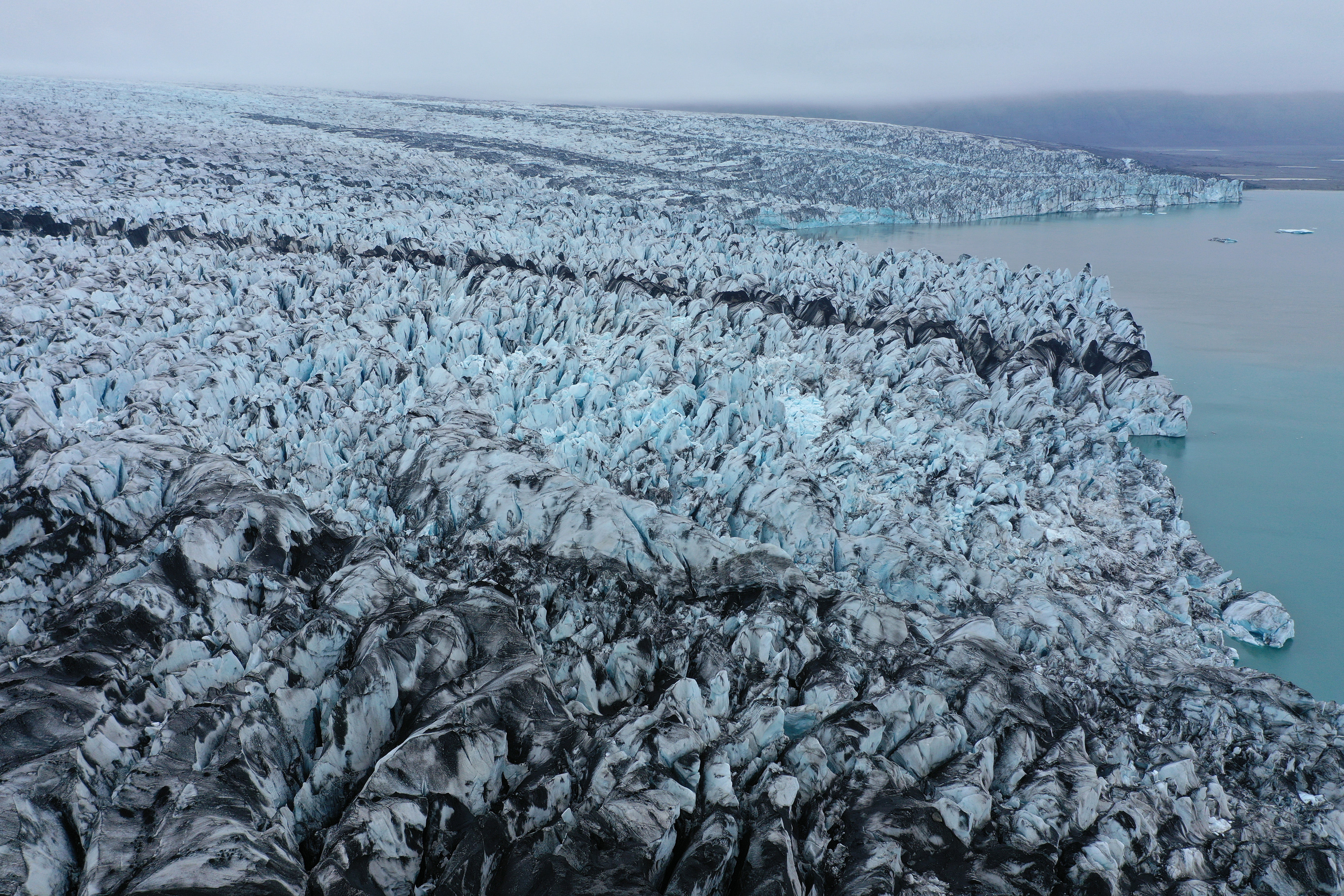 File: In this aerial view receding Breidamerkurjokull glacier, portions of it black with volcanic sand and stones, ends at Jokulsarlon lake on 15 August 2021 near Hof, Iceland