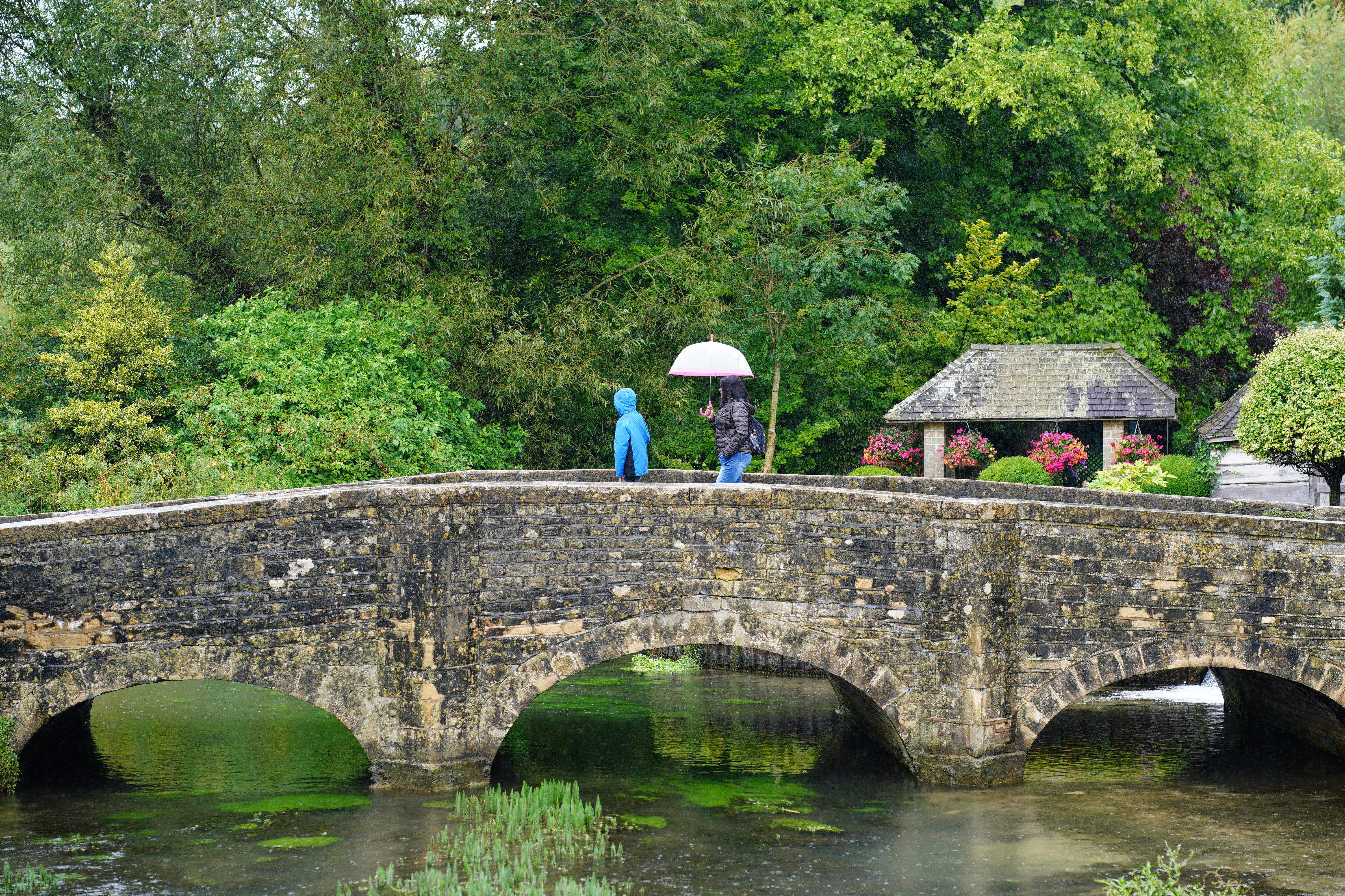 Visitors with umbrellas in the rainy weather at Bibury village in Gloucestershire. Picture date: Saturday August 24, 2024. (Ben Birchall/PA)