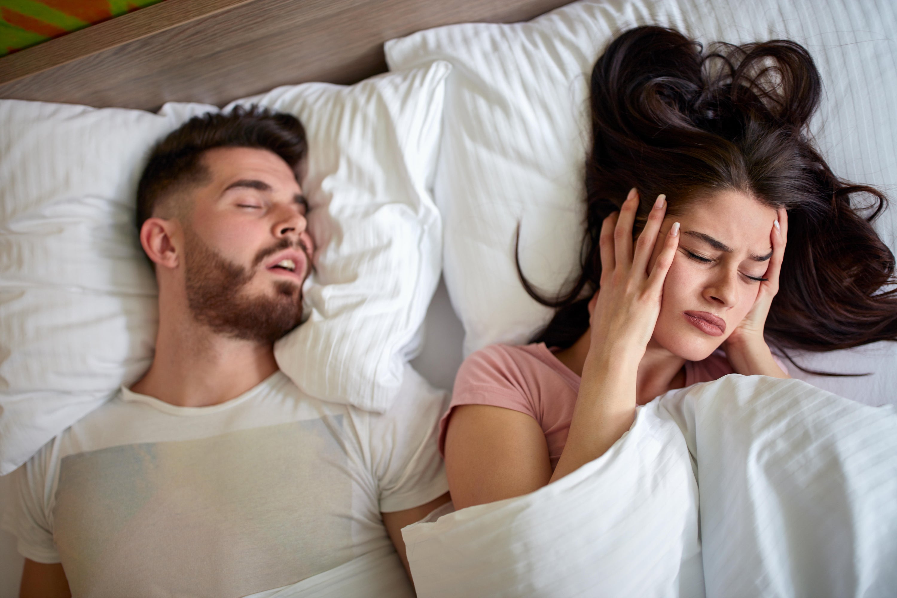 A woman struggling to sleep covering her ears while her partner snores next to her in bed