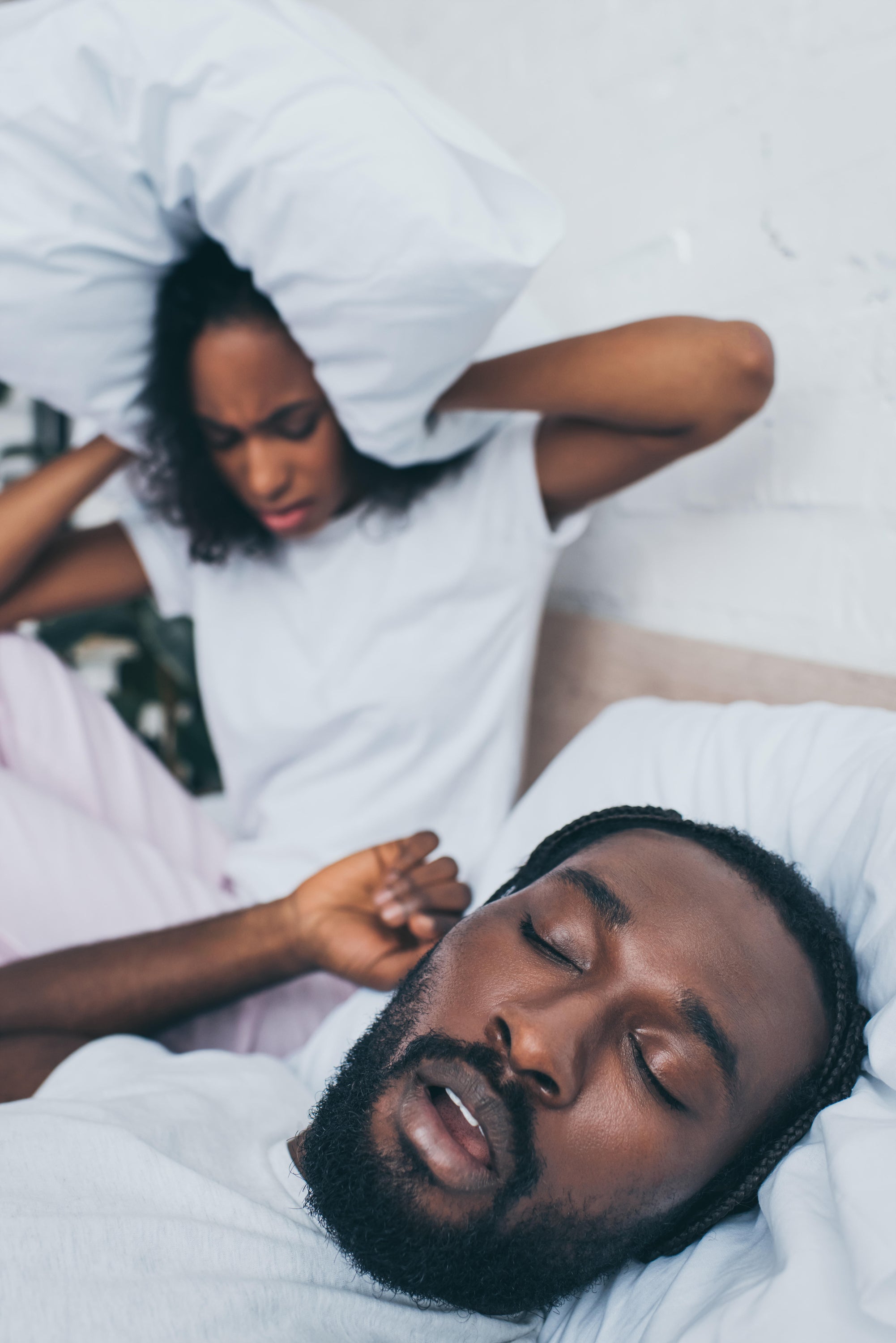 A woman sat up in bed covering her ears with a pillow while her husband snores next to her