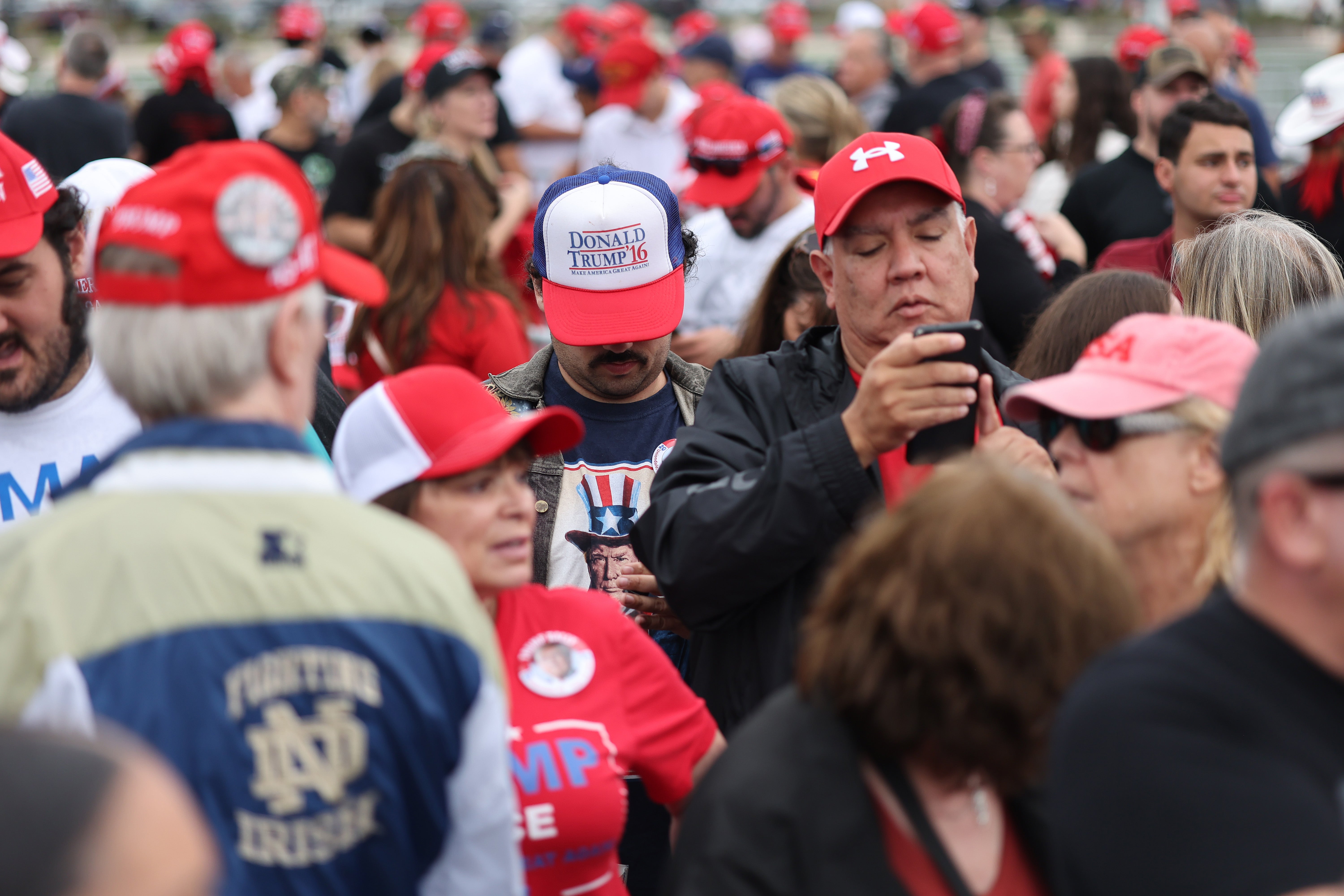 A sea of Trump supporters in red hats outside his rally. One man shows off his long-time support of the former president with a Trump hat from his 2016 presidential run