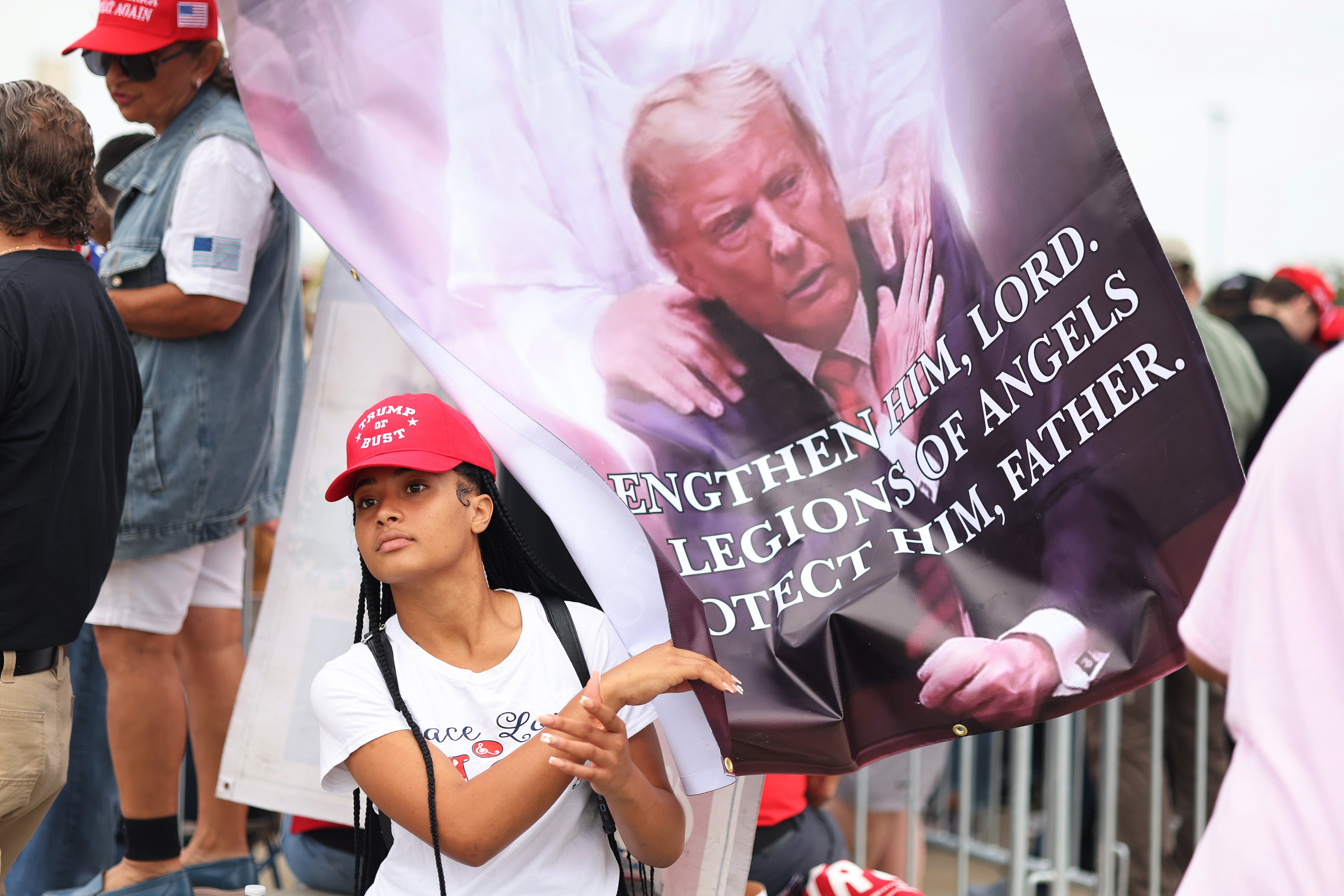 A young Trump fan is nearly engulfed by a huge banner depicting Trump reaching out to touch the hand of Jesus Christ. The deity is pictured clasping Trump’s shoulders. The words “strengthen him Lord” and “legions of angels protect him, Father” are visible on the banner.