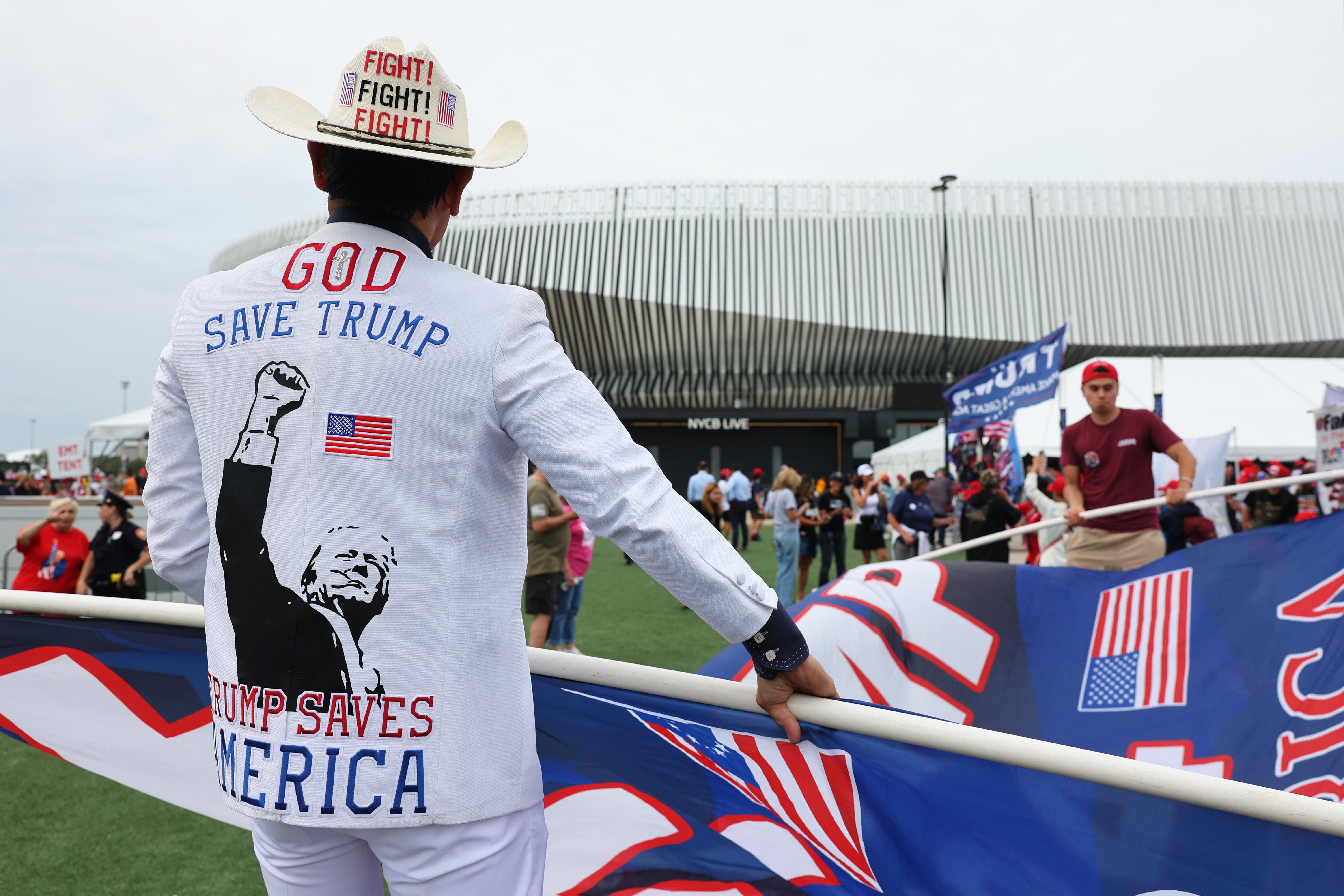 A man decked out in a cowboy hat and a jacket sporting the iconic post-assassination attempt photo of Trump with the words “God saves Trump, Trump saves America” across the back helps another man unfurl a massive flag during a campaign rally