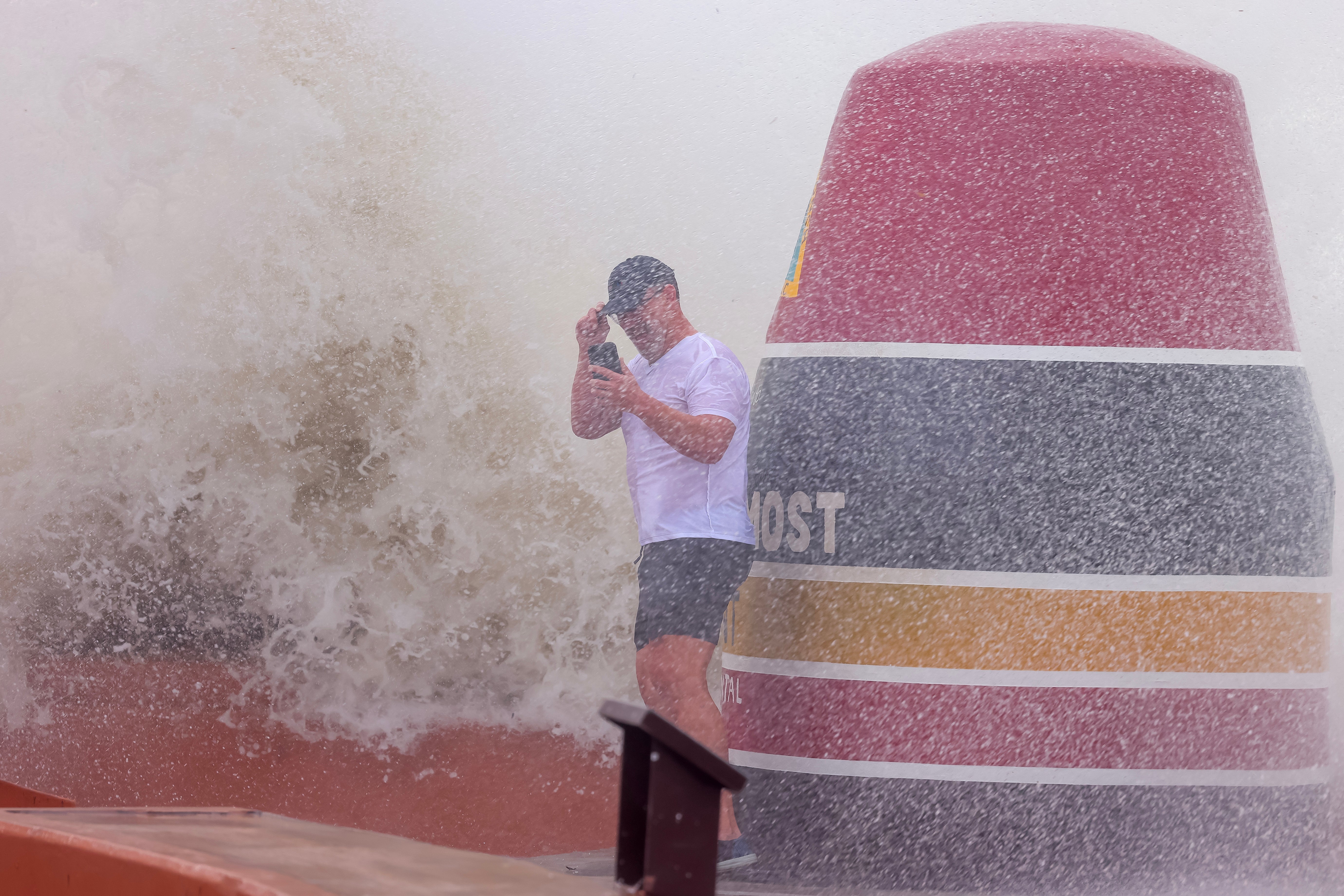 A man holds onto his hat as ocean spray from choppy Atlantic waves nearly surround him at the 'Southernmost Point' buoy in Key West on Thursday