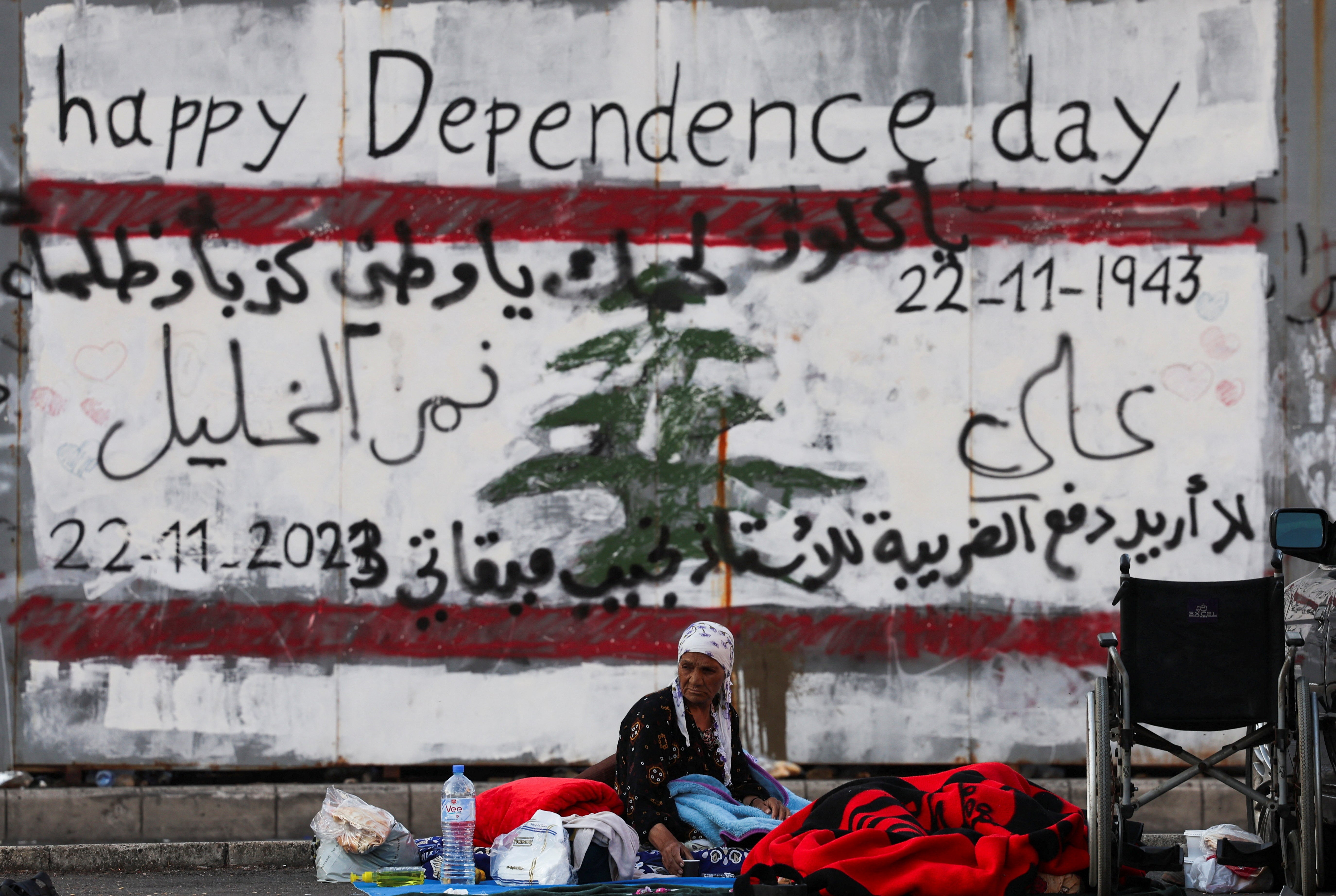 An elderly woman sits with a bottle of water and other belongings in front of Lebanon’s graffitied national flag