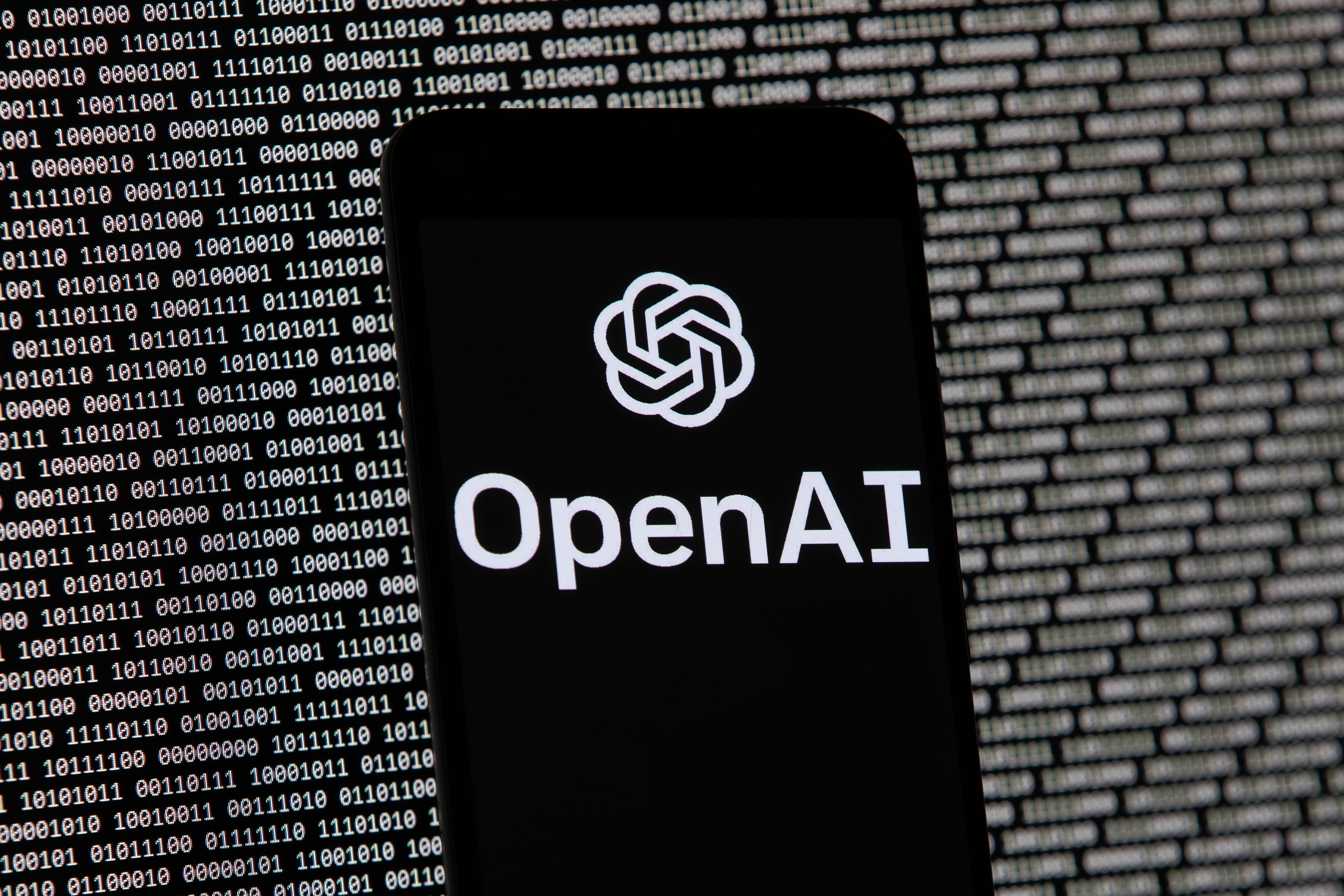 The OpenAI logo appears on a mobile phone in front of a computer screen with random binary data, 9 March, 2023, in Boston