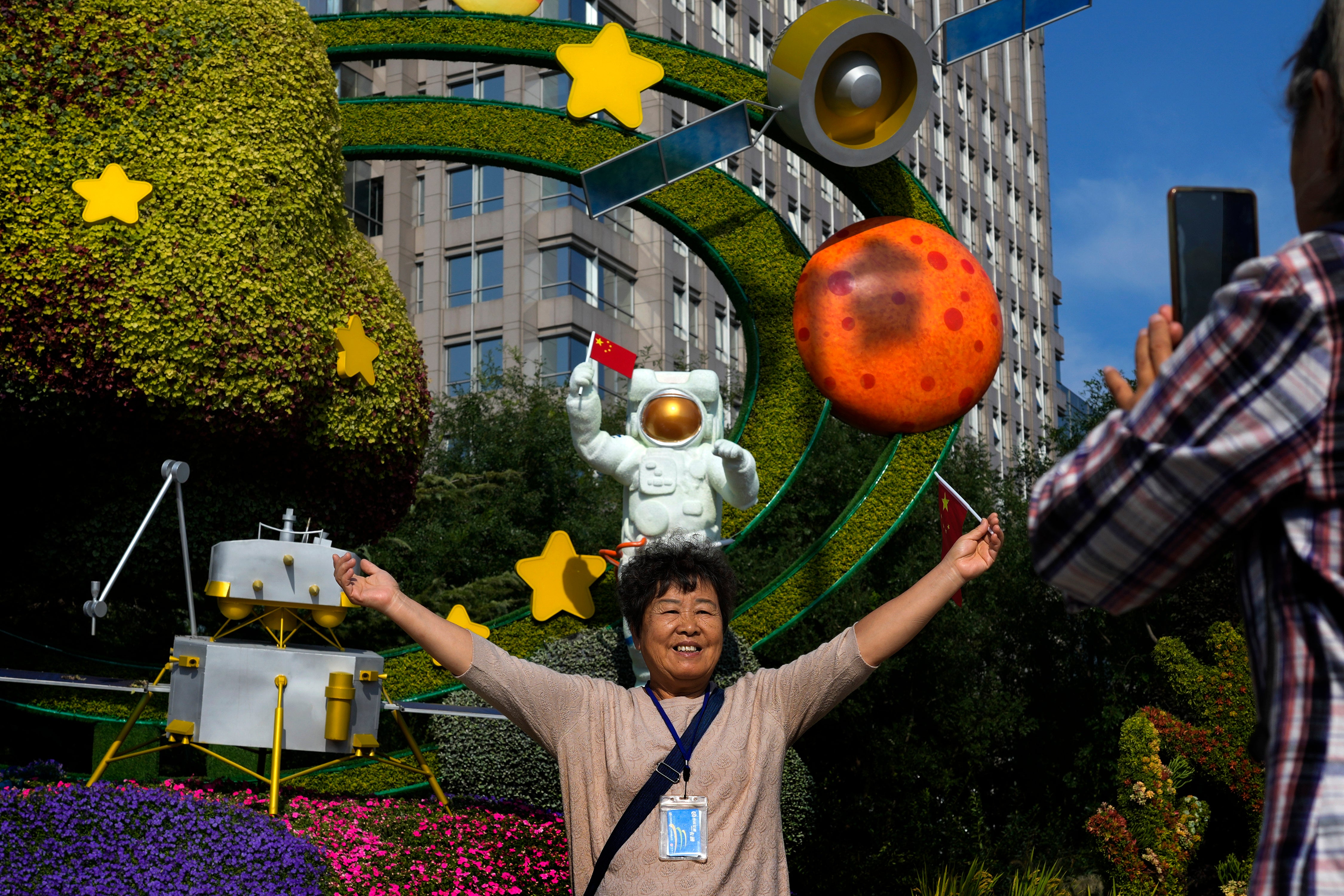 <p>A woman holds a national flag near a floral display featuring a theme of China Space Mission for the upcoming National Day celebration, in Beijing, on 23 September 2024</p>