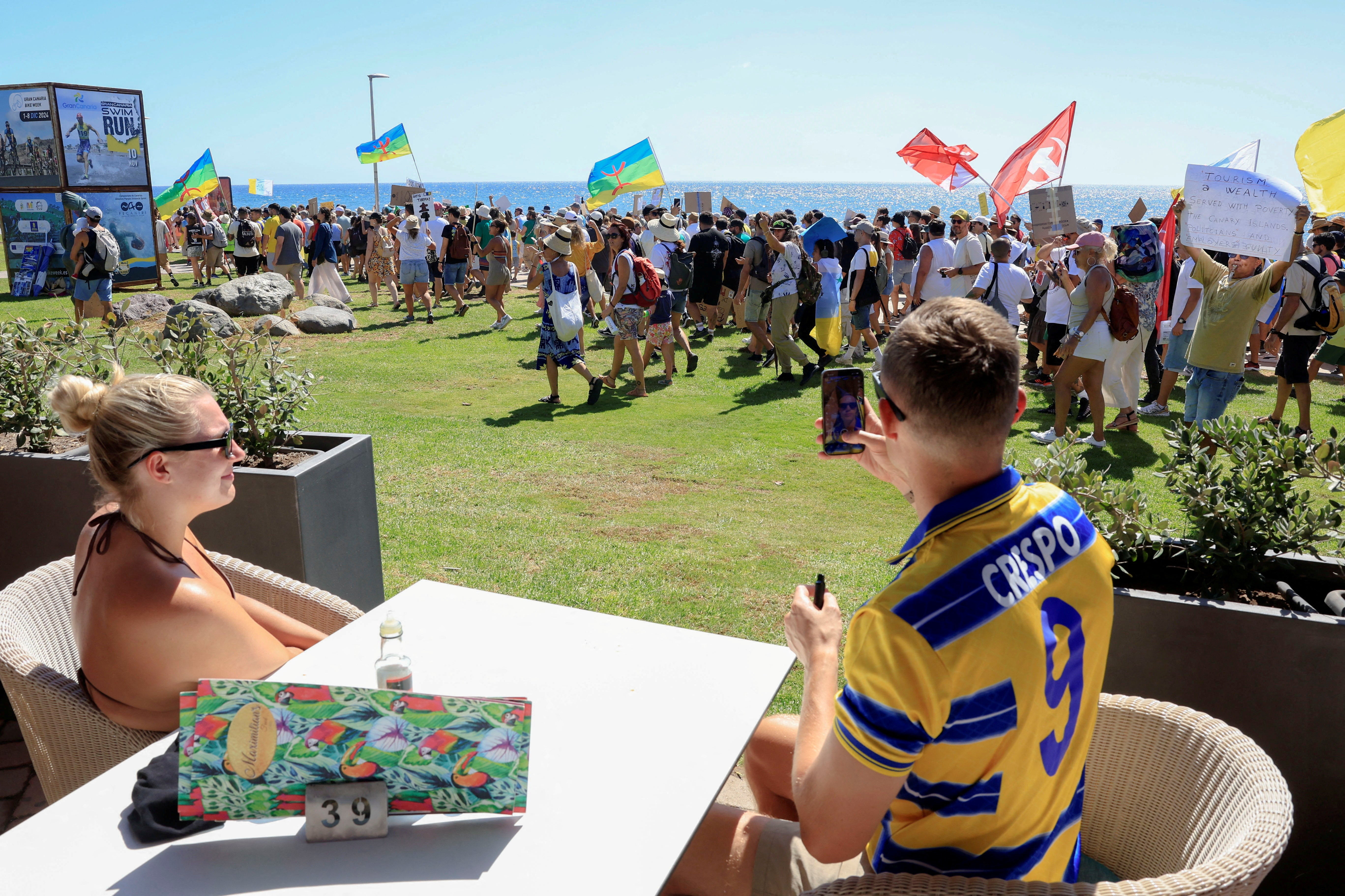 A couple of tourists sitting on a terrace watch a demonstration for a change in the tourism model in the Canary Islands on the island of Gran Canaria, Spain