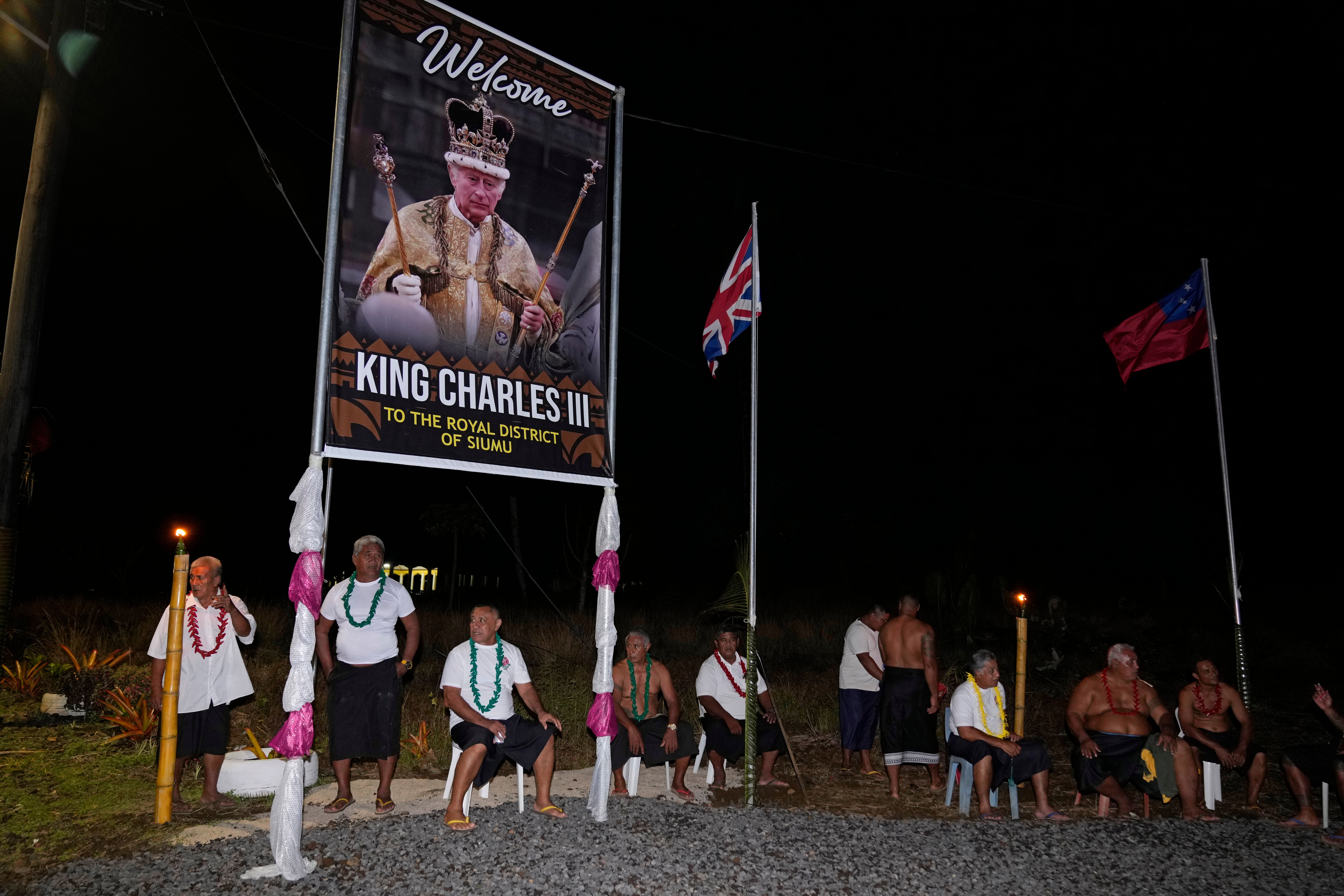 People sit under a portrait of Britain's King Charles III as they wait for his arrival and Queen Camilla in the village of Siumu, Samoa.