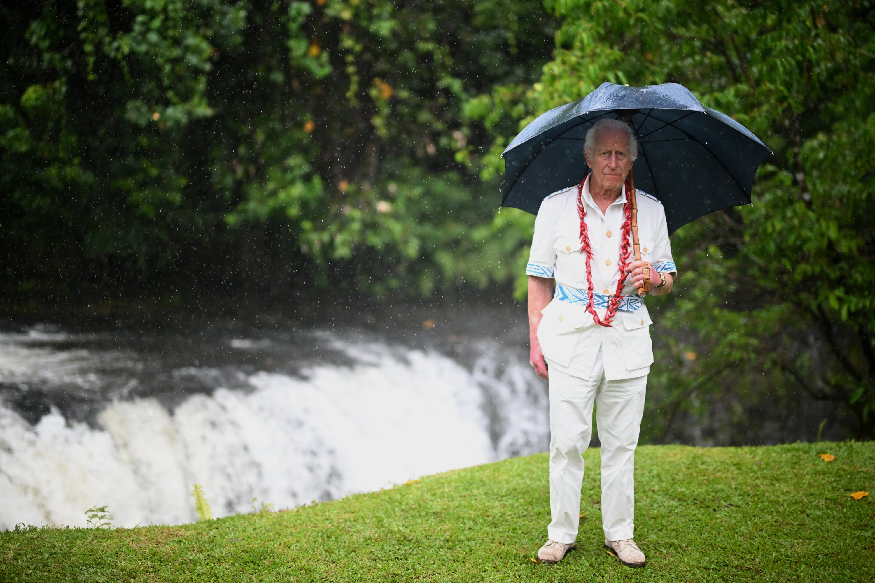 El rey Carlos se encuentra junto a una cascada mientras usa un paraguas para protegerse de la lluvia durante su visita al Parque Nacional O Le Pupu'Pue.