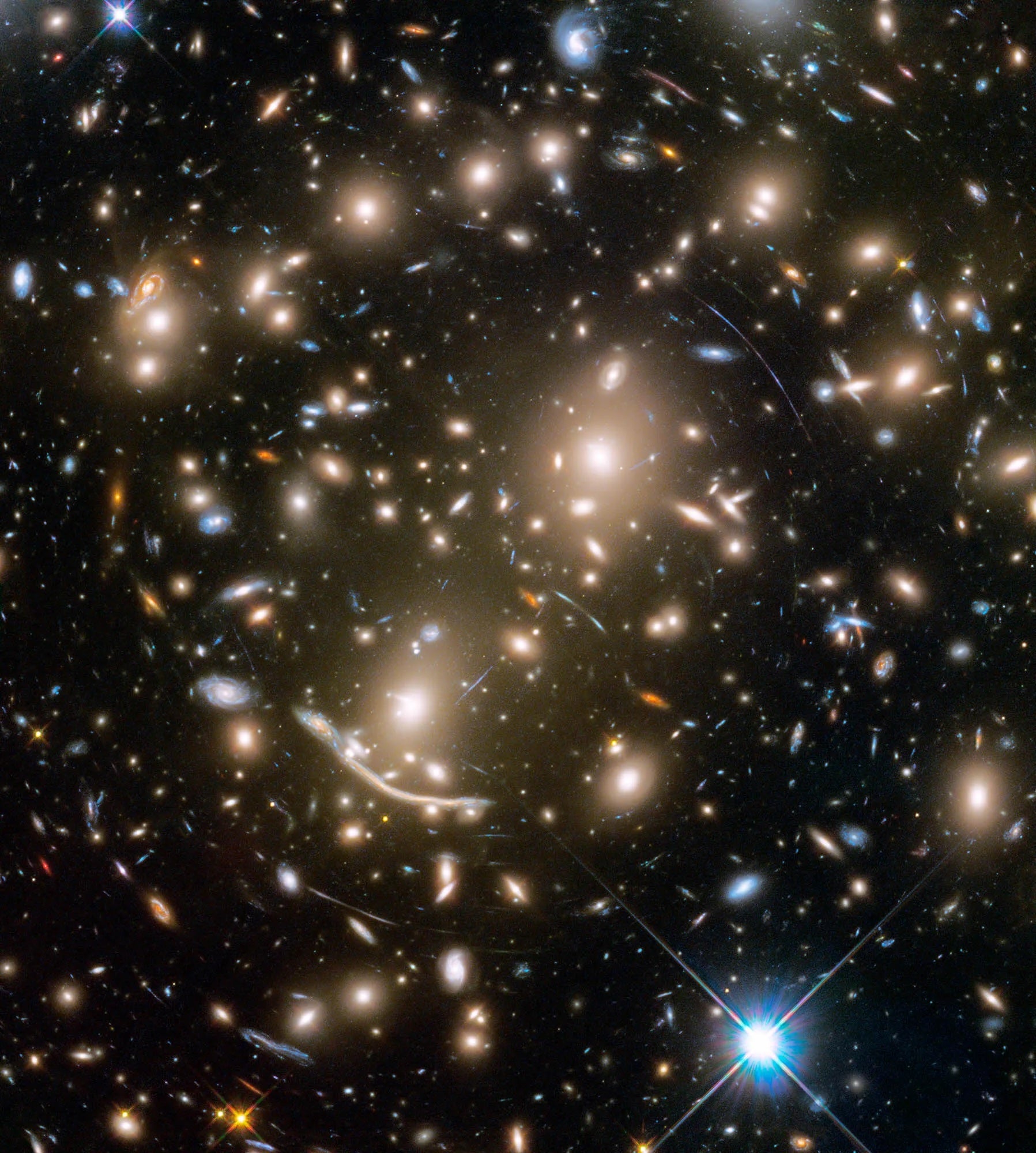 Among the galaxies in this Hubble Space Telescope image are mysterious arcs of blue light. The arcs are distorted images of remote galaxies. The collective gravity of all the normal and dark matter trapped inside the galaxy cluster warps space-time, affecting light traveling toward Earth