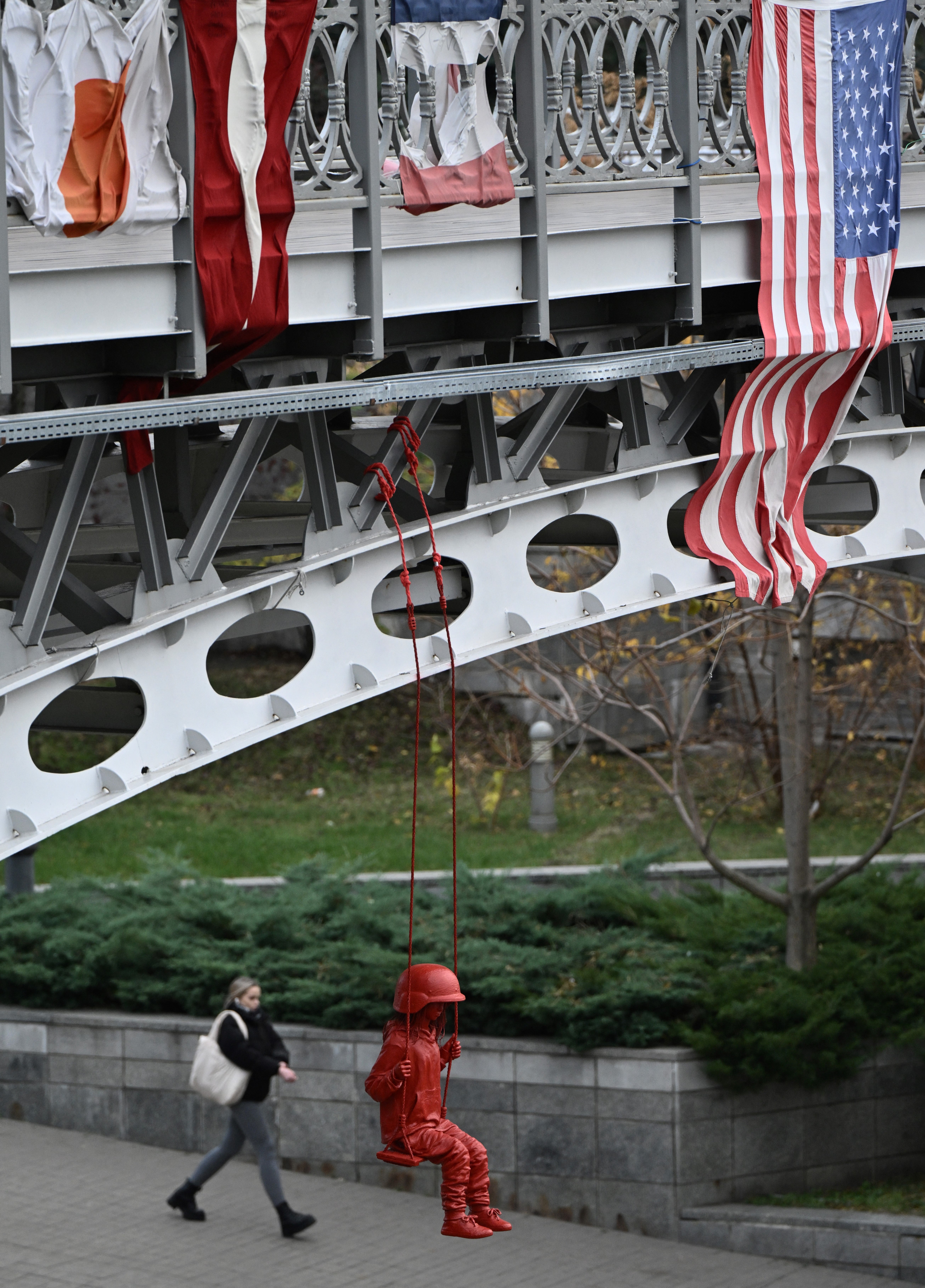 American flag hung off a bridge next to an art installation by French street artist James Colomina called the "Swing" which depicts a little girl wearing an oversized military helmet as she swings above the Alley of the Heroes of the Heavenly Hundred at Independence Square in Kyiv