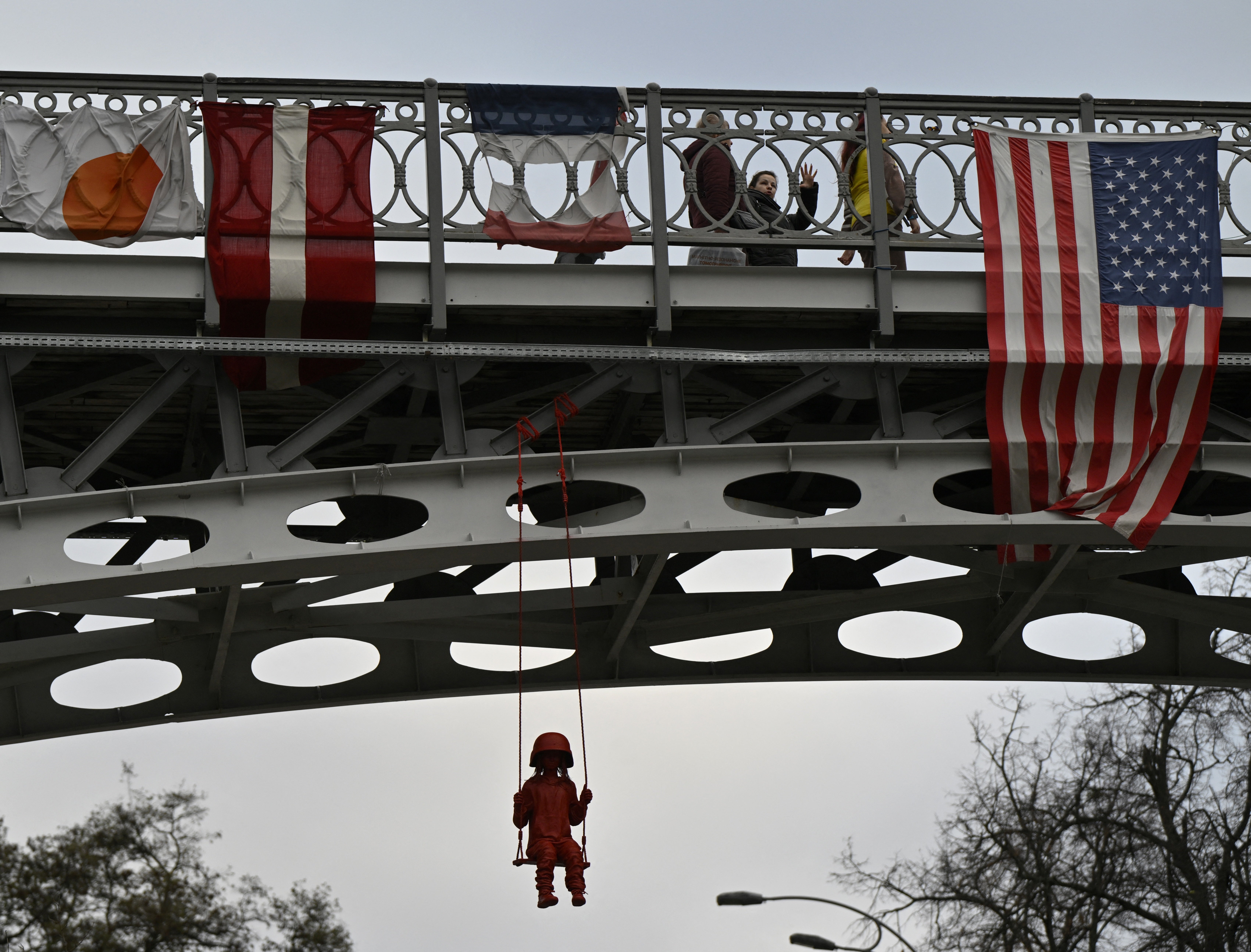 People walk past an American flag hung off a bridge next to an art installation by French street artist James Colomina called the “Swing” which depicts a little girl wearing an oversized military helmet as she swings above the Alley of the Heroes of the Heavenly Hundred at Independence Square in Kyiv