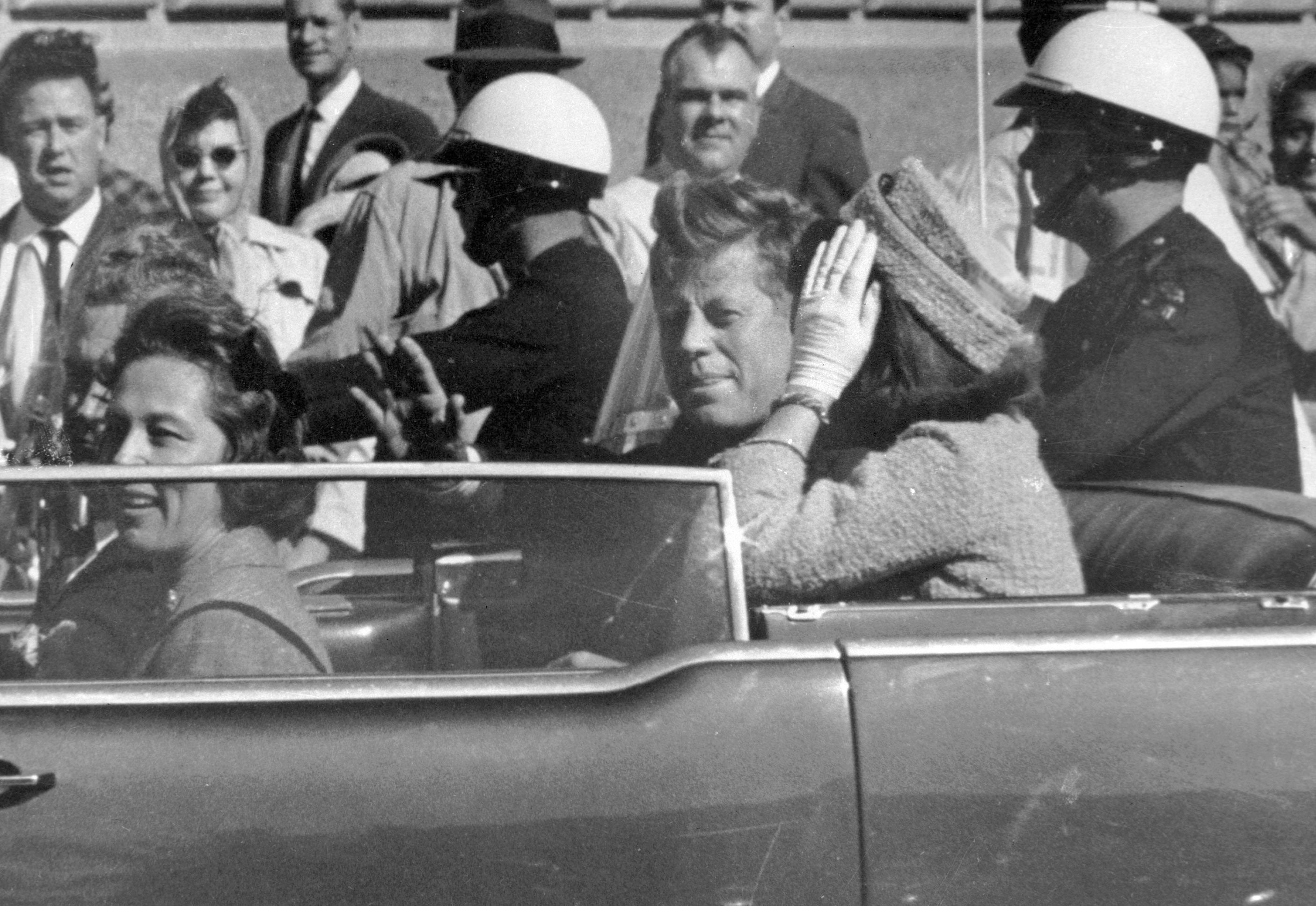 President John F. Kennedy waves from his car in a motorcade approximately one minute before he was shot