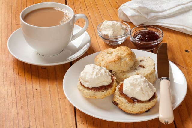 <p>A plate of three scones with jam and cream next to a cup of tea with a saucer</p>