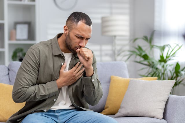 <p>A young Black man coughs as he sits on a couch</p>