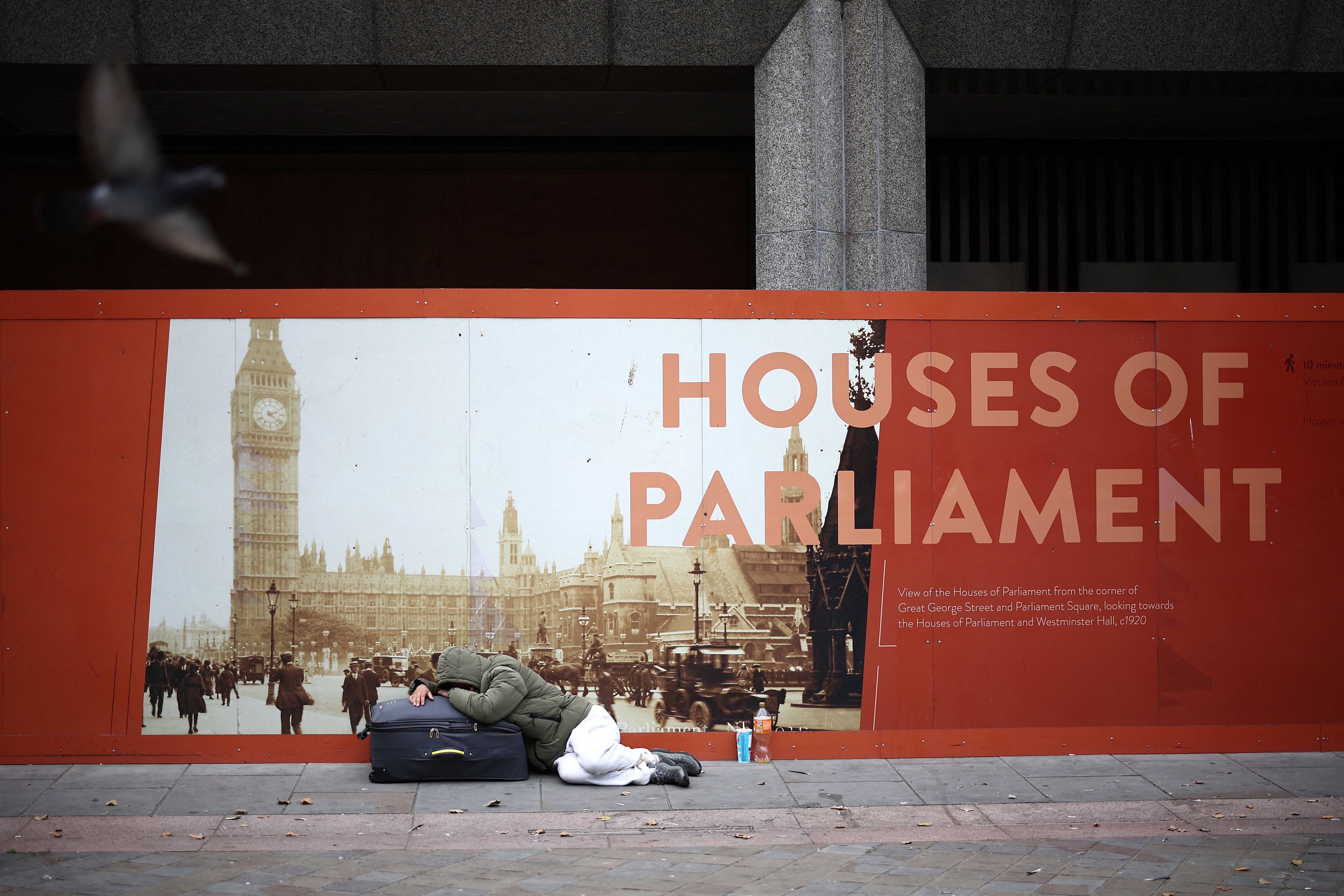 A rough sleeper lies on a suitcase in front of an advertisement board showing the Houses of Parliament in central London on September 15, 2024