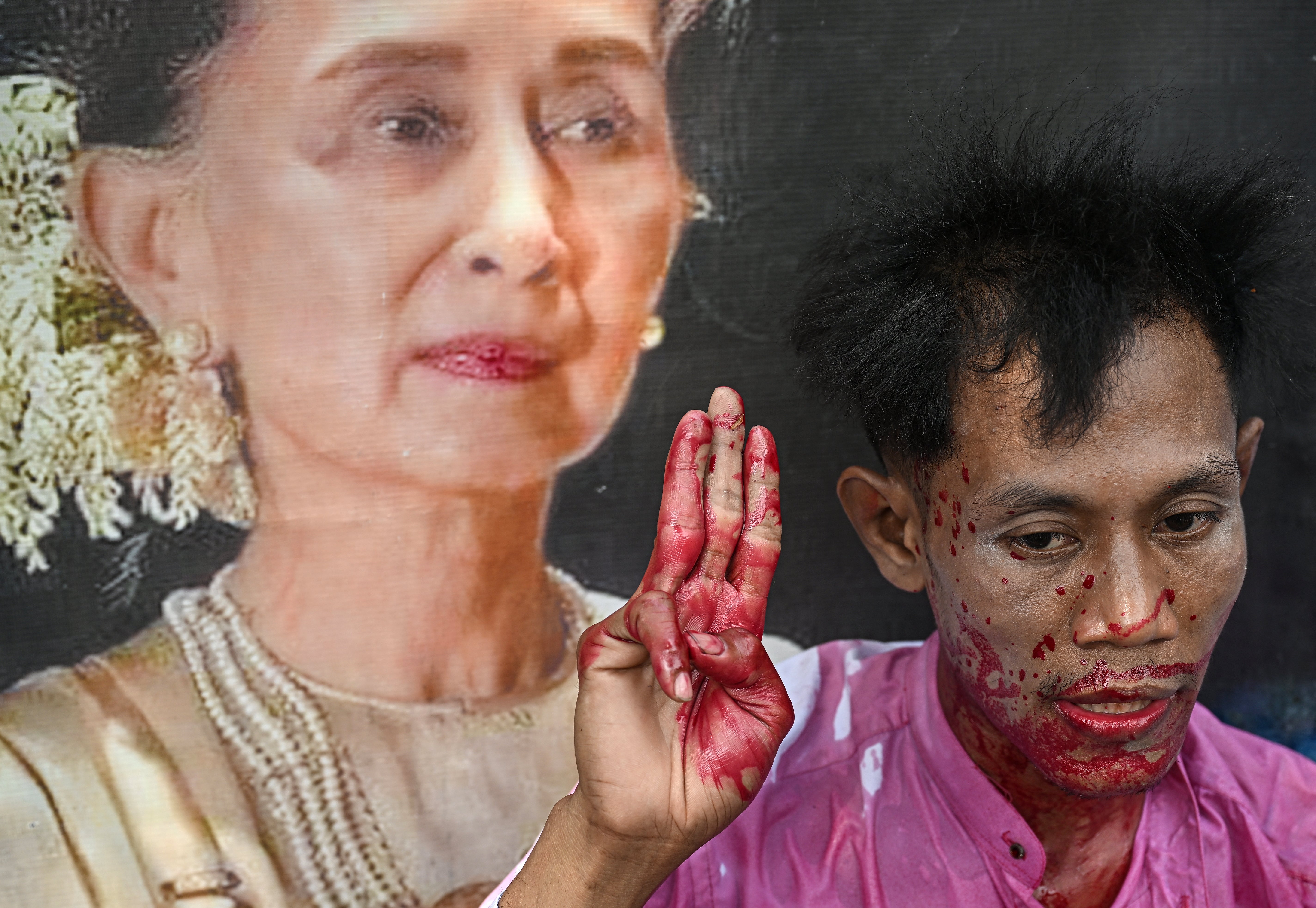 A protester covered in fake blood next to an image of detained leader Aung San Suu Kyi during a demonstration outside the UN office in Bangkok