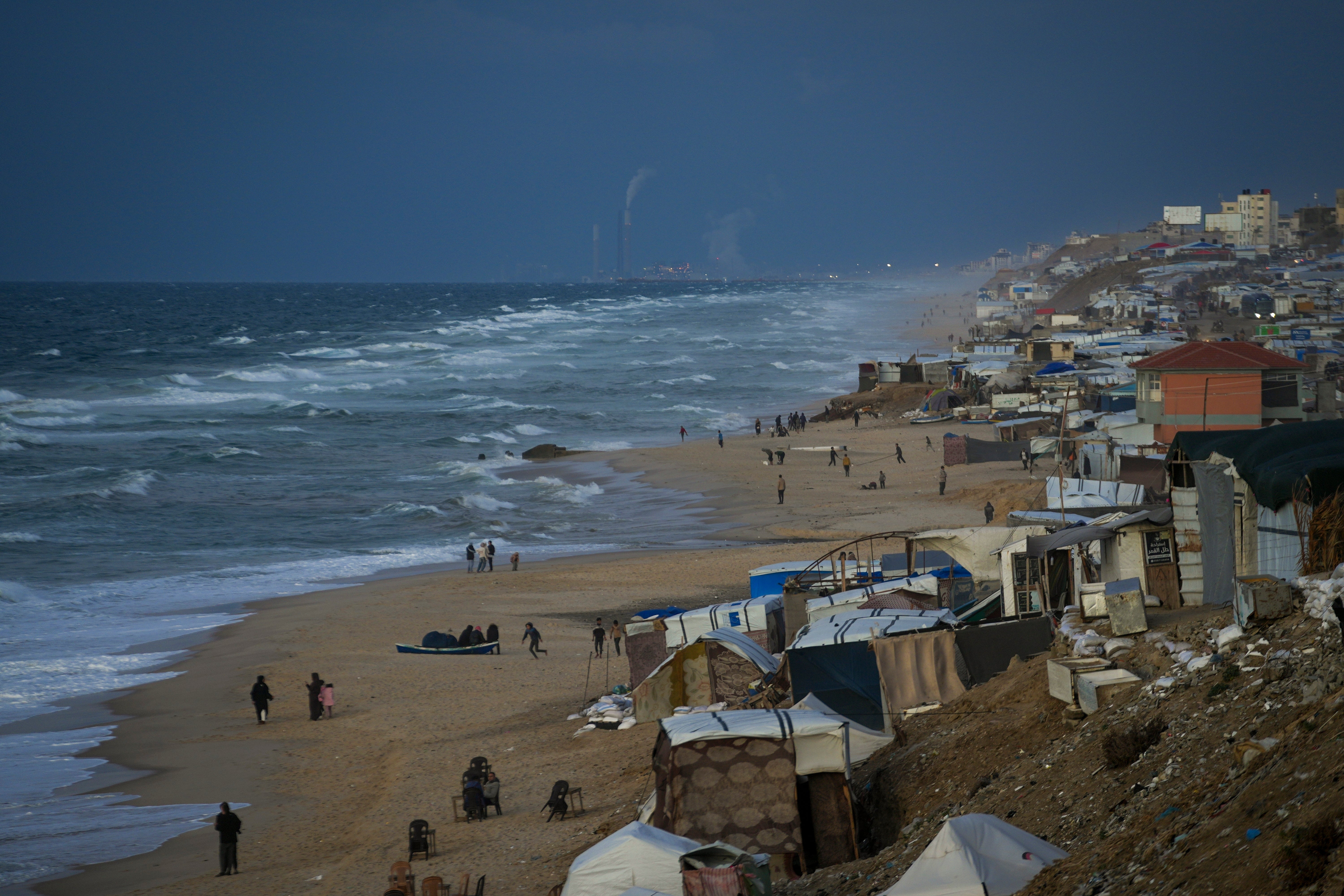 People walk along the beach next to a tent refugee camp for displaced Palestinians in Deir al-Balah, central Gaza Strip, Monday, December 30, 2024