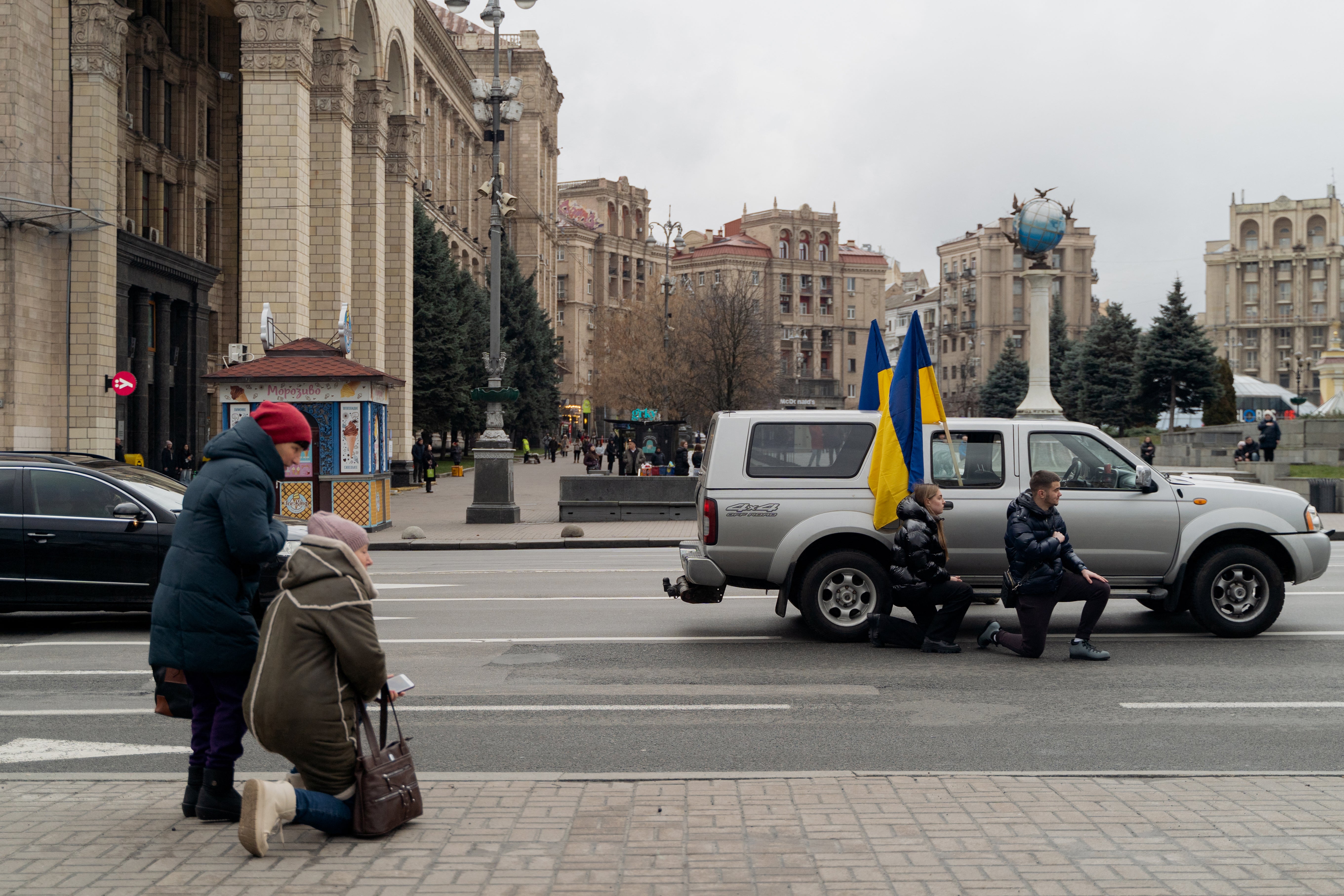 People crossing a street stop as they kneel to honour a fallen Ukranian soldier in Kyiv