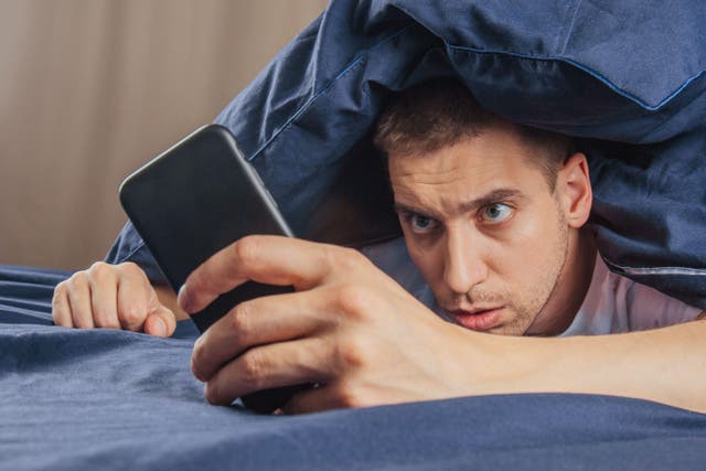 <p>A shocked man laying in bed under a blue duvet looking at his phone</p>