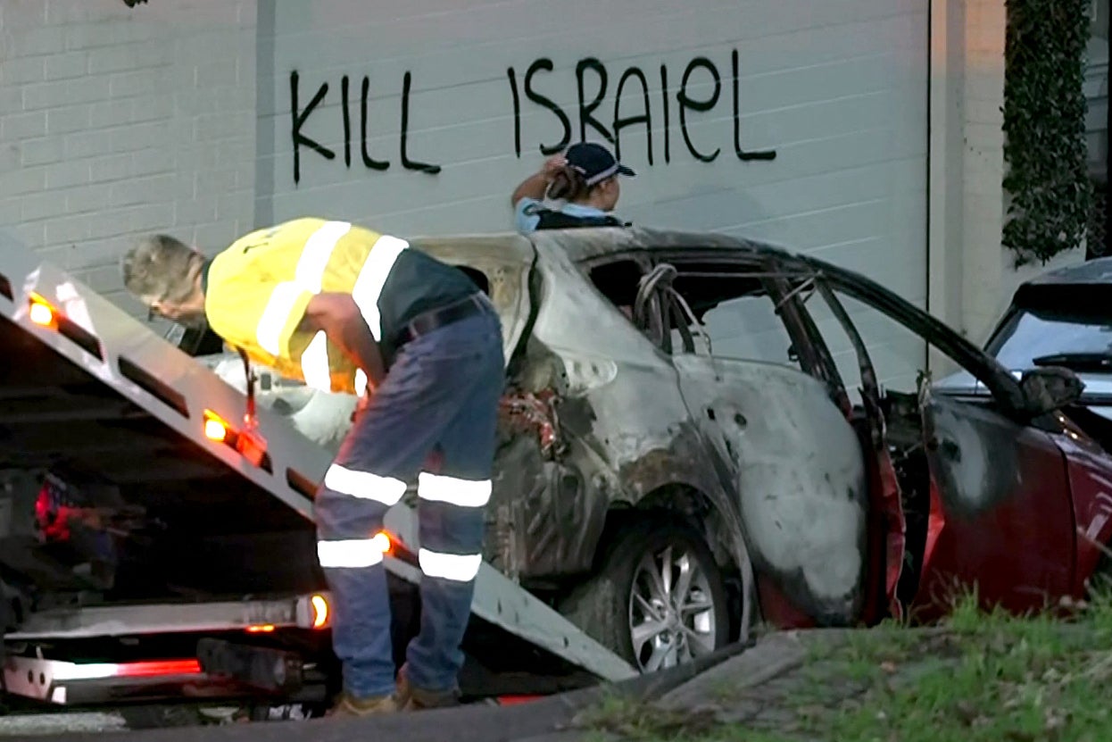 This frame grab taken from video footage provided by the Australian Broadcast Corporation (ABC) on December 11, 2024 shows a police officer walking past a torched car being removed in front of anti-Israel graffiti