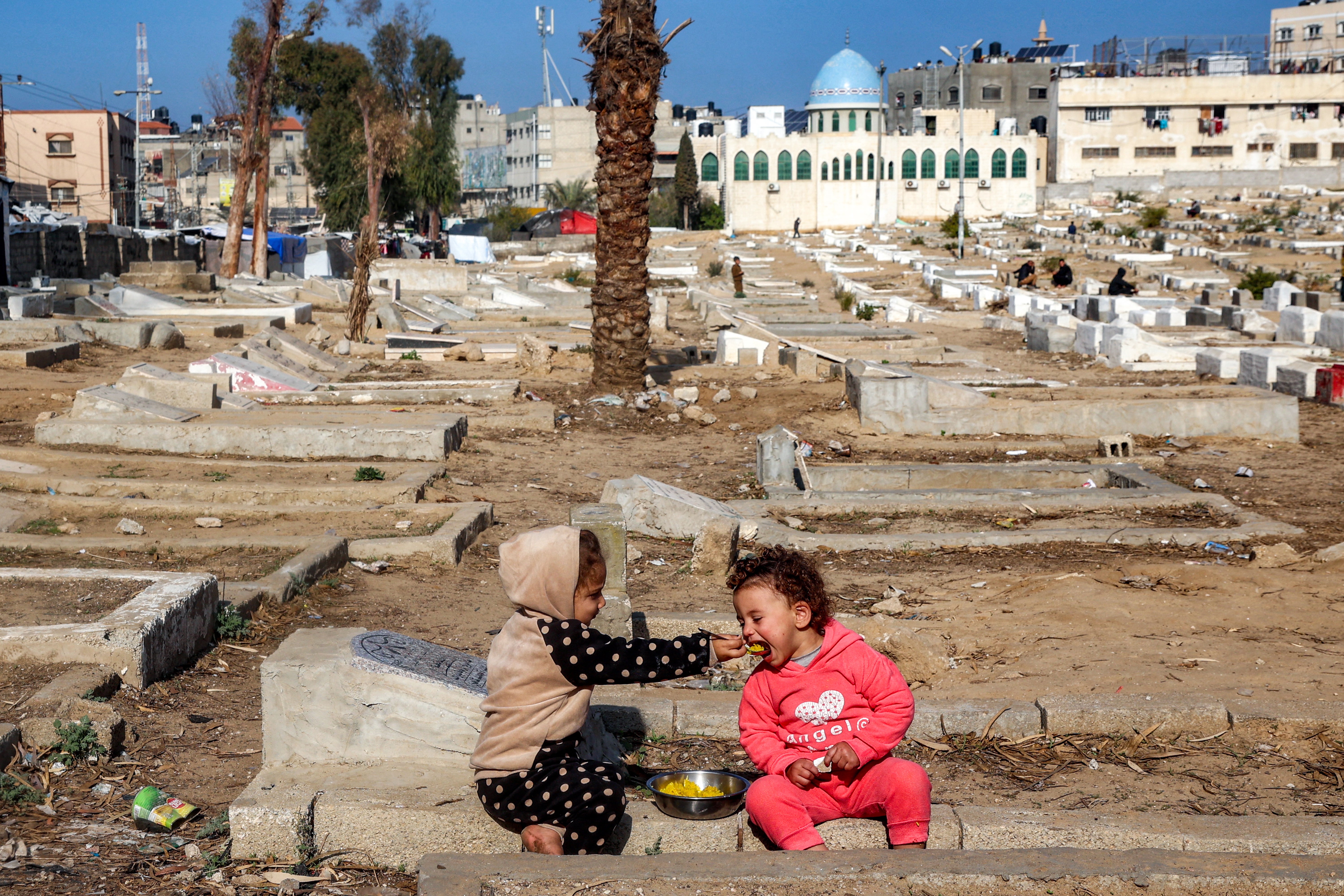 A child feeds another a spoonful of food as they sit atop graves at a cemetery where families displaced by conflict are taking shelter in Deir el-Balah in the central Gaza Strip