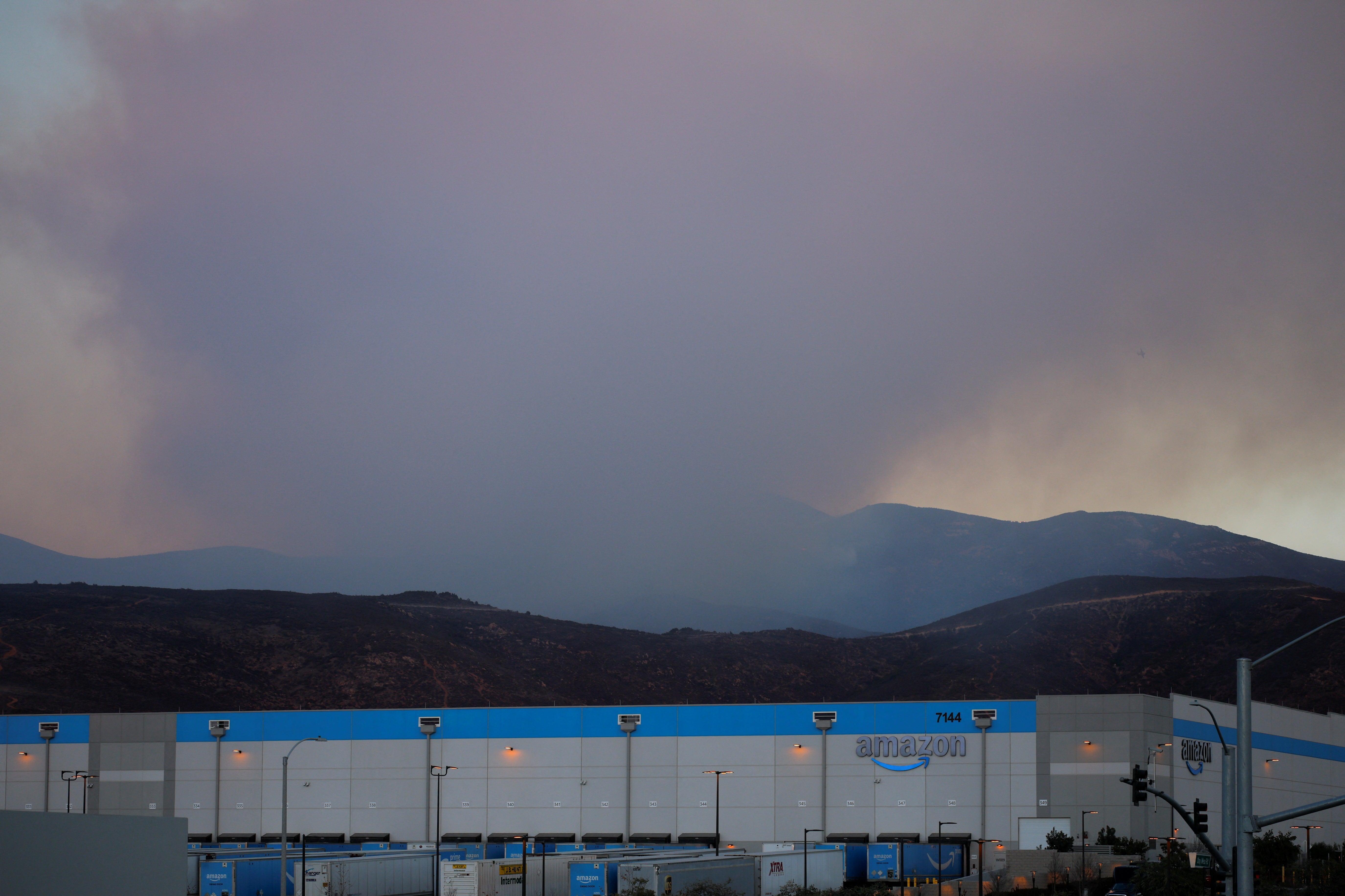 Smoke rises from the hills behind an Amazon warehouse as firefighters battle the Border 2 fire next to the Mexican border, in the hills east of the Otay Mesa neighborhood of San Diego, California U.S, January 23, 2025. REUTERS/Mike Blake