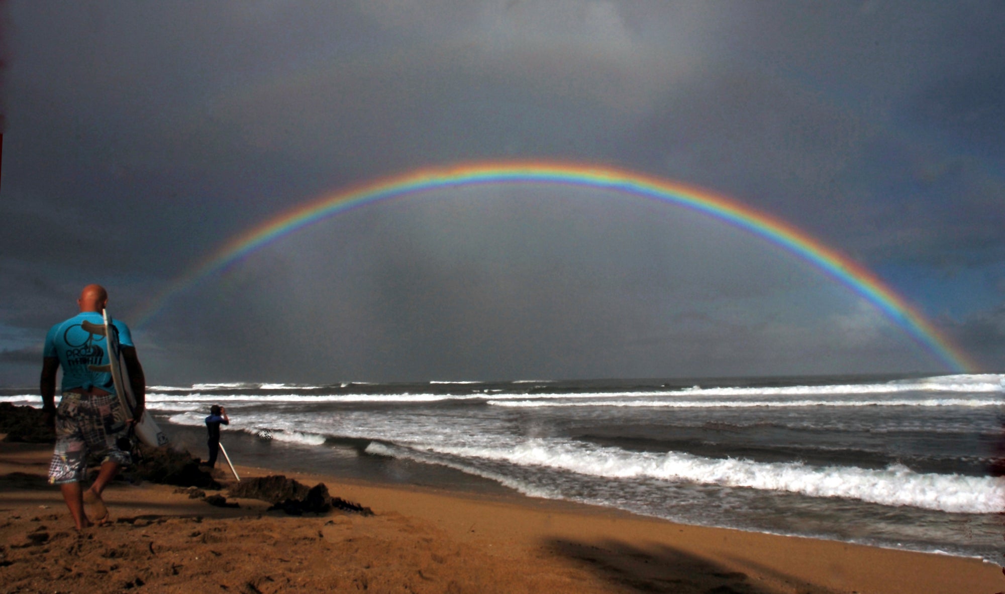 Los surfistas profesionales se dirigen al agua mientras un gran arco iris cruza el océano en Hawaii