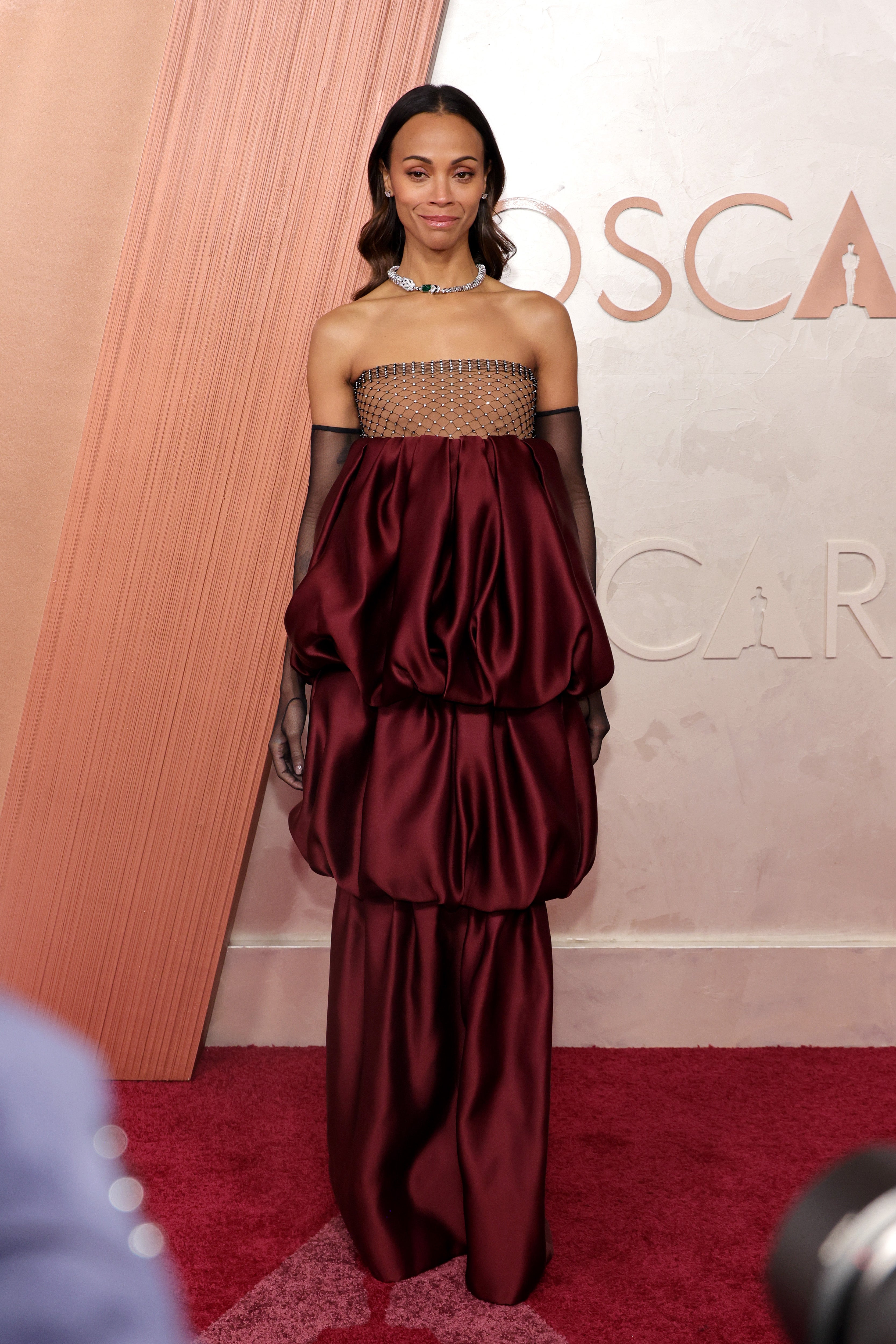 Zoe Saldaña posed on the Oscars red carpet outside the Dolby Theater in a three-tiered, wine-colored gown with a strapless black net neckline and matching opera gloves