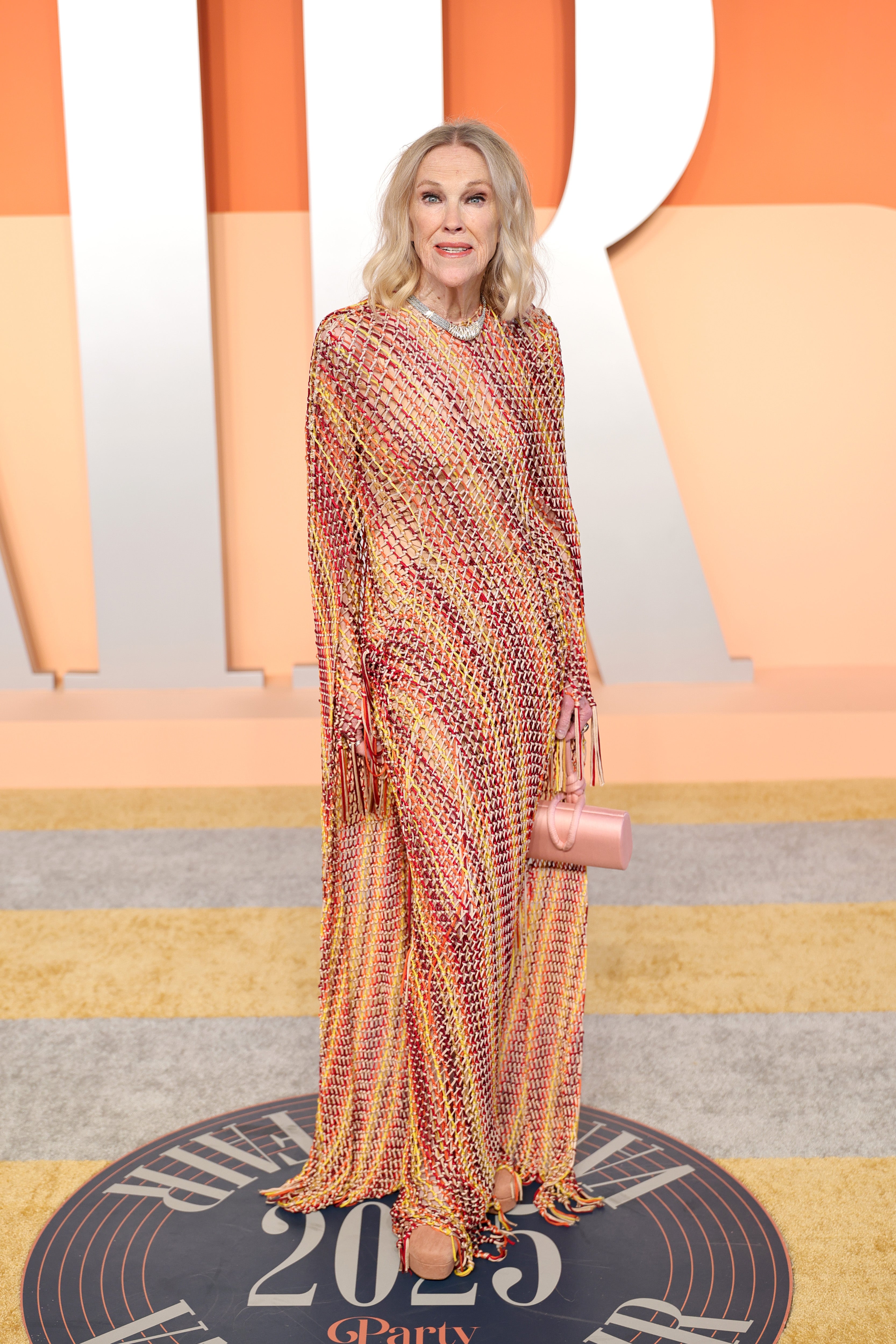 Catherine O’Hara matched the warm, sunset-hued backdrop on the after-party red carpet in a woven long-sleeved gown