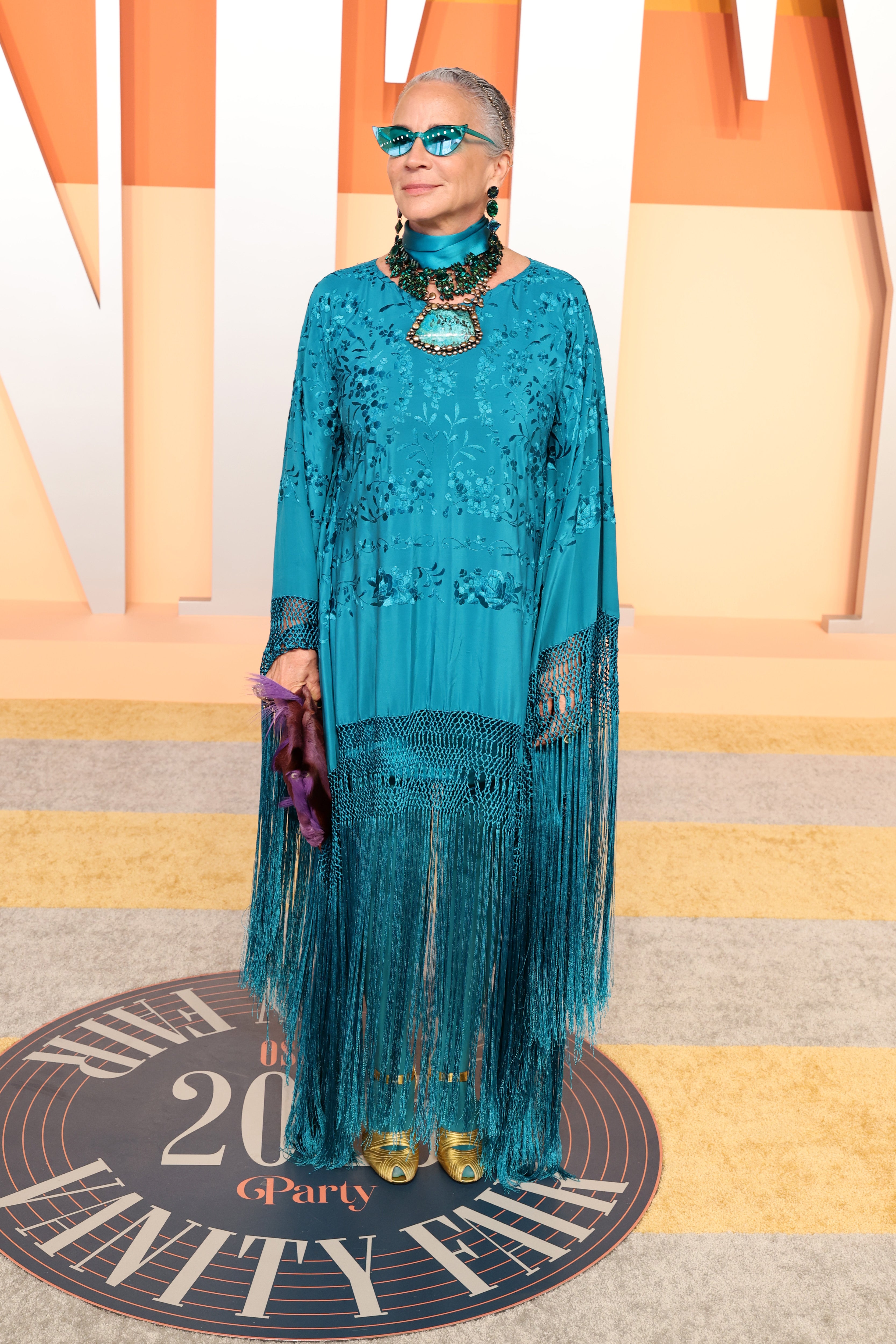 Lisa Eisner posed in a teal-toned tunic with long fringe trim, a turquoise statement-making choker necklace, and matching shades