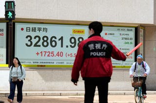 Monitors, back, show Japan's Nikkei 225 index at a securities firm as a member of the Tokyo Metropolitan Police directs traffic and pedestrians