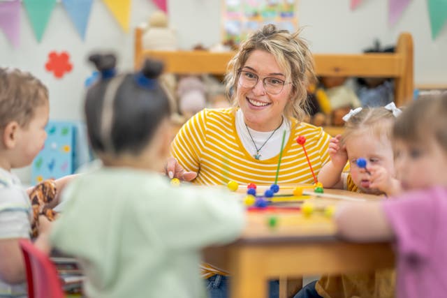 <p>A small group of preschool children sit with their teacher at a table as they learn through play.  They are each dressed casually and are focused on the activity.</p>