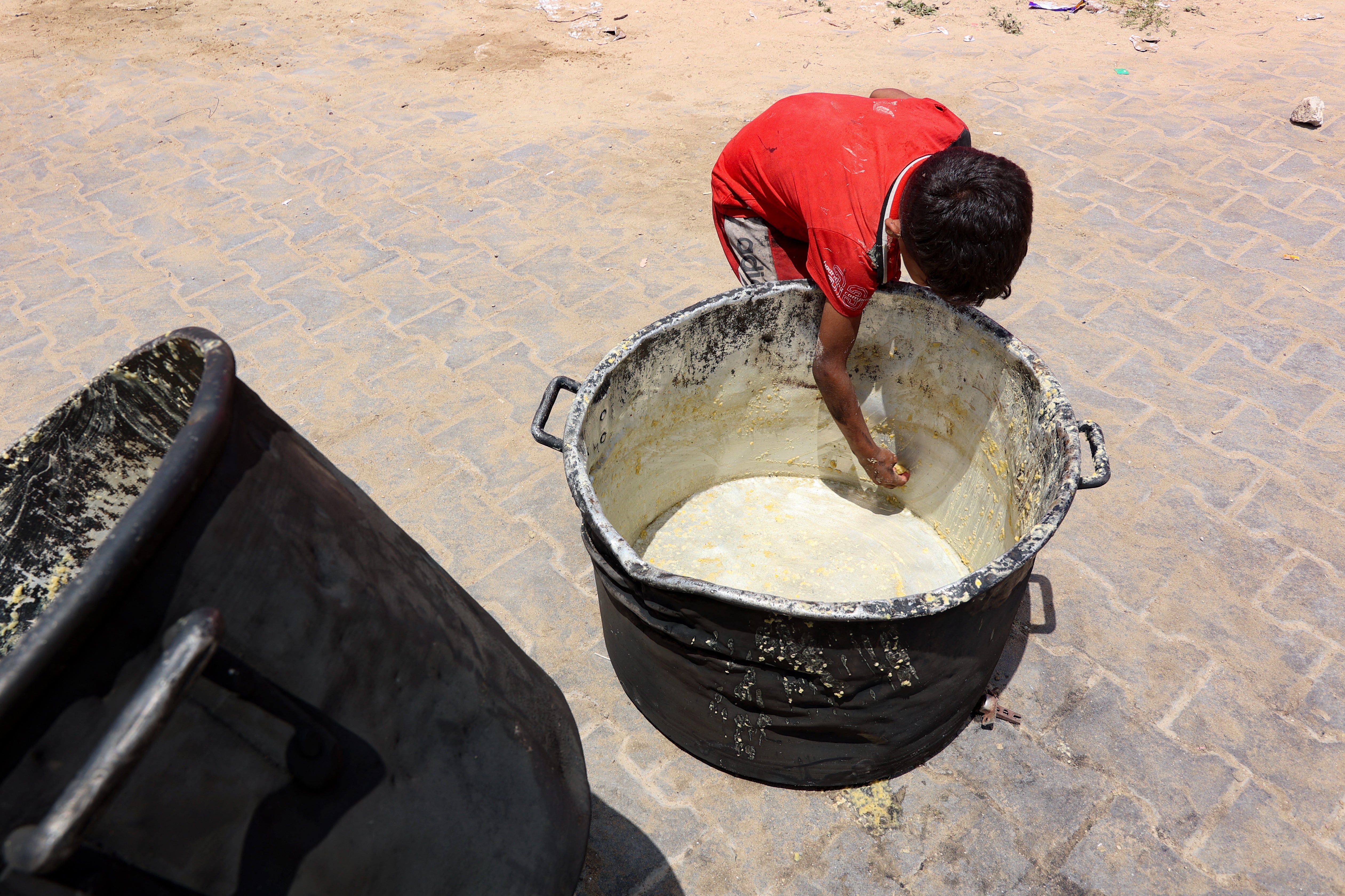 A Palestinian boy scrapes off bits of lentil soup remaining in a cooking pot, in front of a food distribution point at a displacement camp