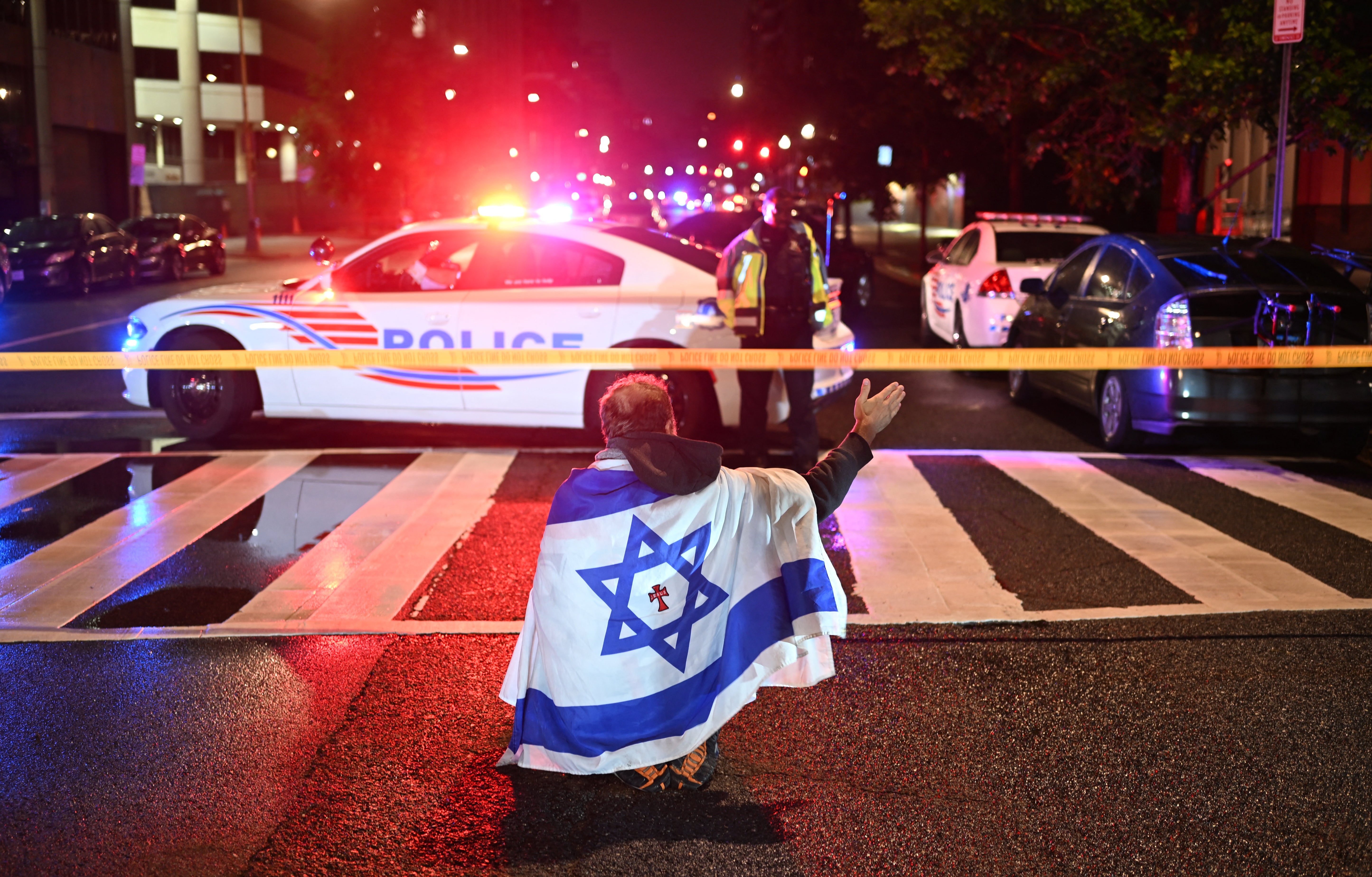 A man draped in the Israeli flag kneels behind the police cordon outside the Capital Jewish Museum
