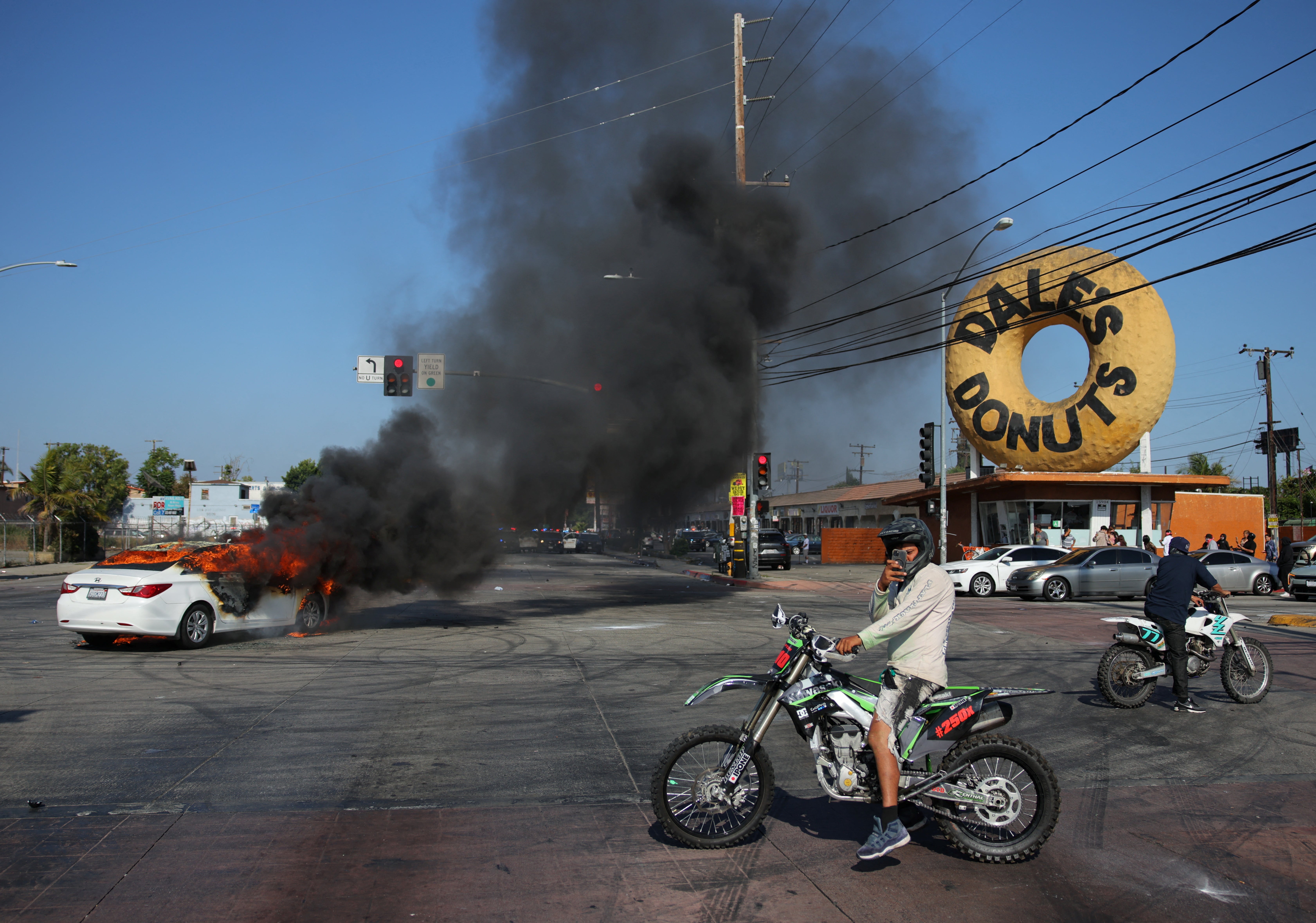 People ride motorbikes next to a car burning in flames during a standoff between police and protesters following multiple detentions by Immigration and Customs Enforcement (ICE), in the Los Angeles County city of Paramount, California, U.S., June 7, 2025