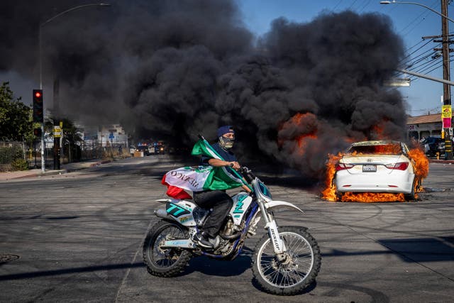 <p>A man on a motorcycle carries a Mexican flag as smoke rises from a burning car on Atlantic Boulevard, during a standoff by protesters and law enforcement, following multiple detentions by Immigration and Customs Enforcement (ICE), in the Los Angeles County city of Paramount, California, U.S., June 7, 2025</p>