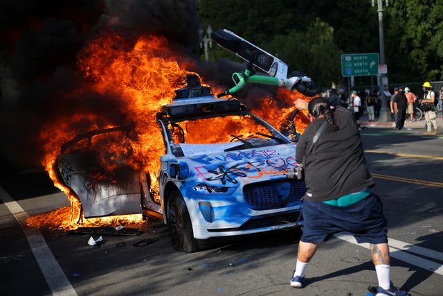 <p>Un manifestante lanza un patinete eléctrico contra un coche en llamas durante una protesta contra las redadas federales de inmigración en el centro de Los Ángeles</p>