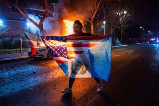 A person displaying multiple flags walks past a burning car during protests