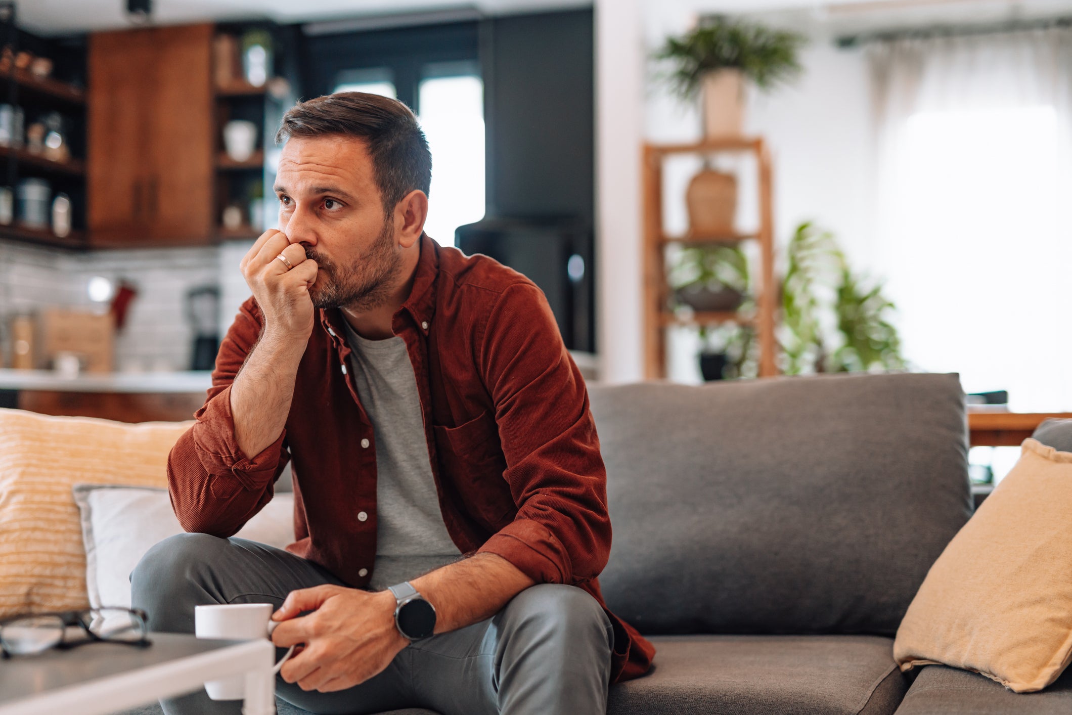 <p>Worried man sitting on the sofa at home, biting fingernails while holding a cup of coffee</p>