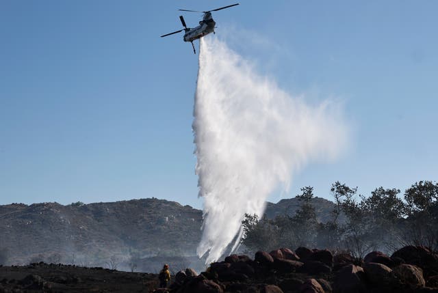 <p>A firefighting helicopter drops water as a firefighter (bottom L) works while the Juniper Fire burns on June 30, 2025</p>